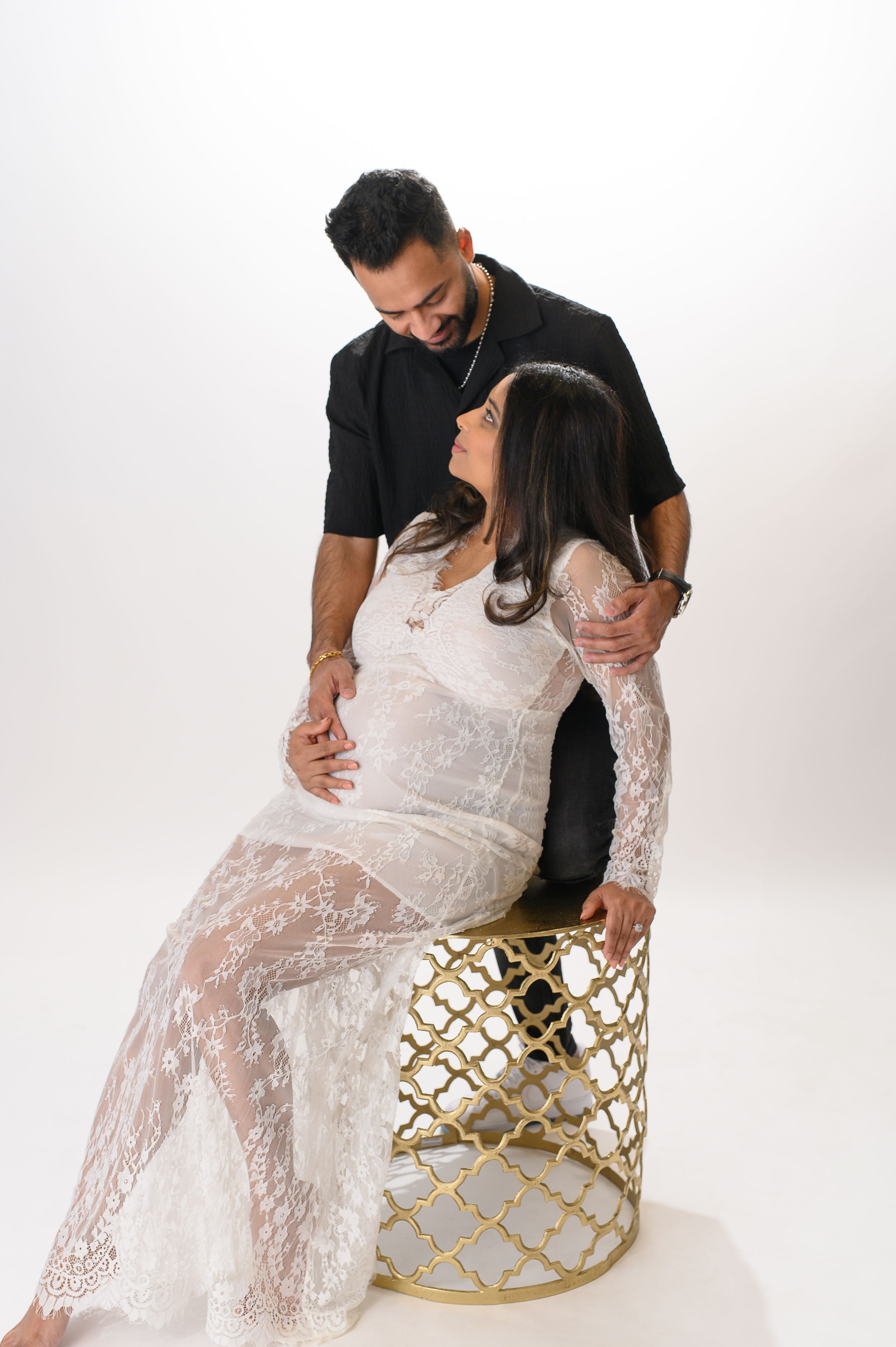 Romantic photo shoot with couple in a white lace dress and black shirt, posing intimately on a gold geometric stool.