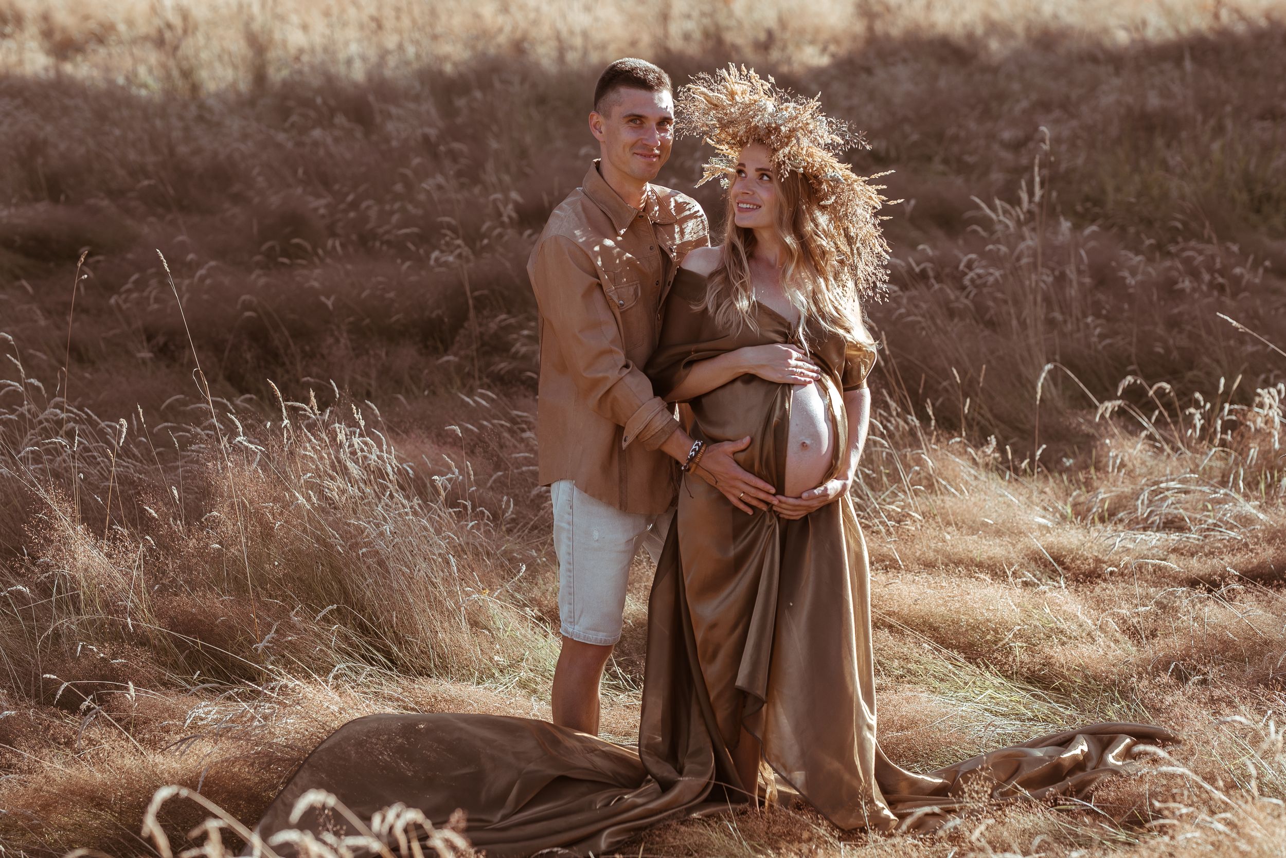 A couple in earth-toned clothing poses together in a sun-drenched field of tall dry grass during a maternity photoshoot.