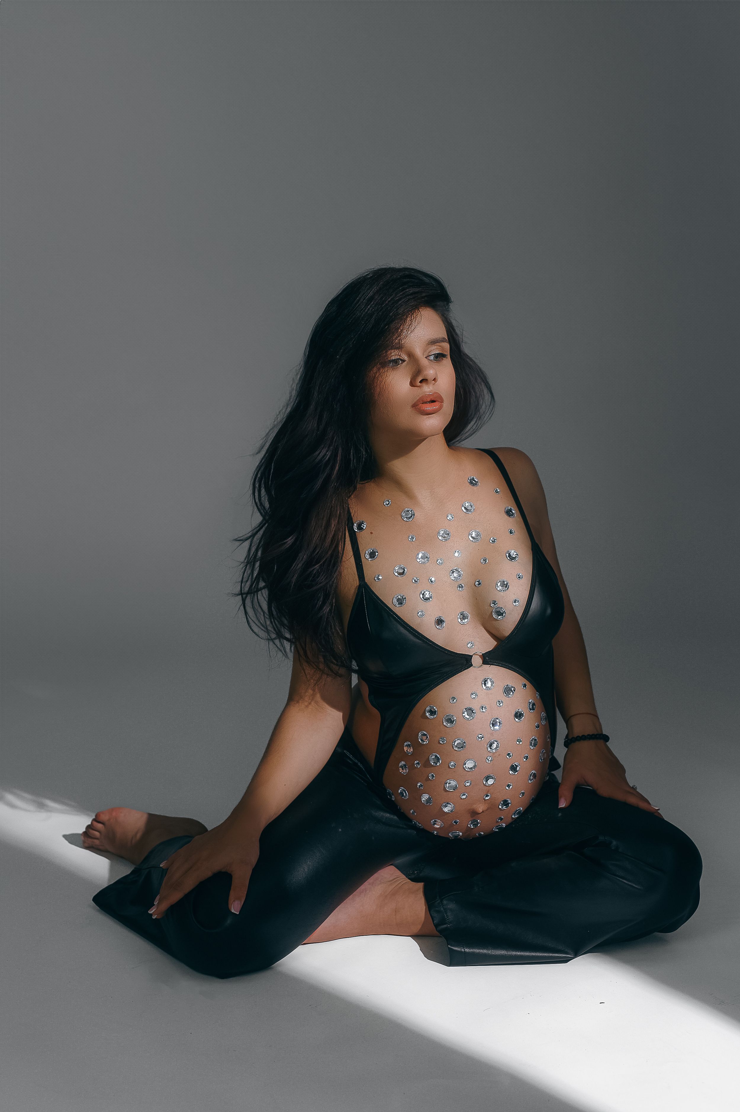 Artistic fashion portrait of model in sheer black dress with jeweled embellishments sitting on studio floor in dramatic lighting.