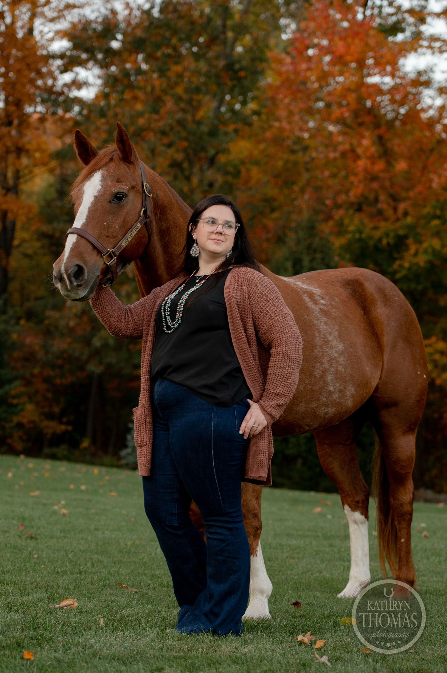 A chestnut horse with white socks stands with a companion on a grassy hill against vibrant autumn foliage.