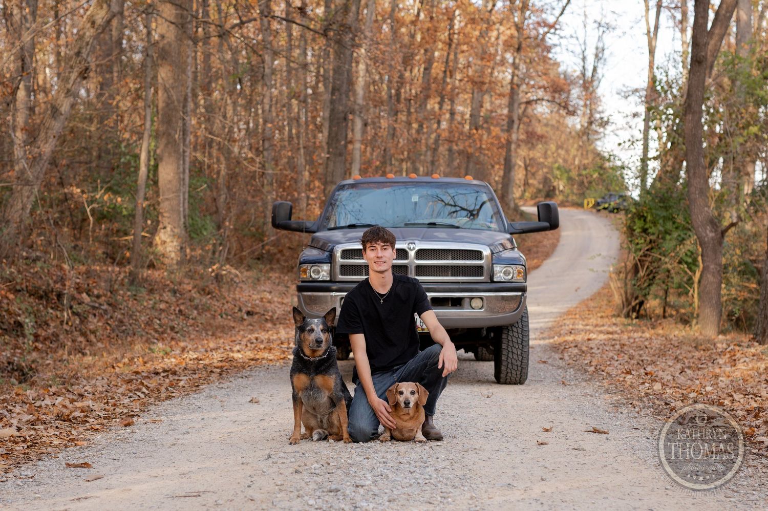 Person kneeling with two dogs beside silver Dodge truck on wooded dirt road.