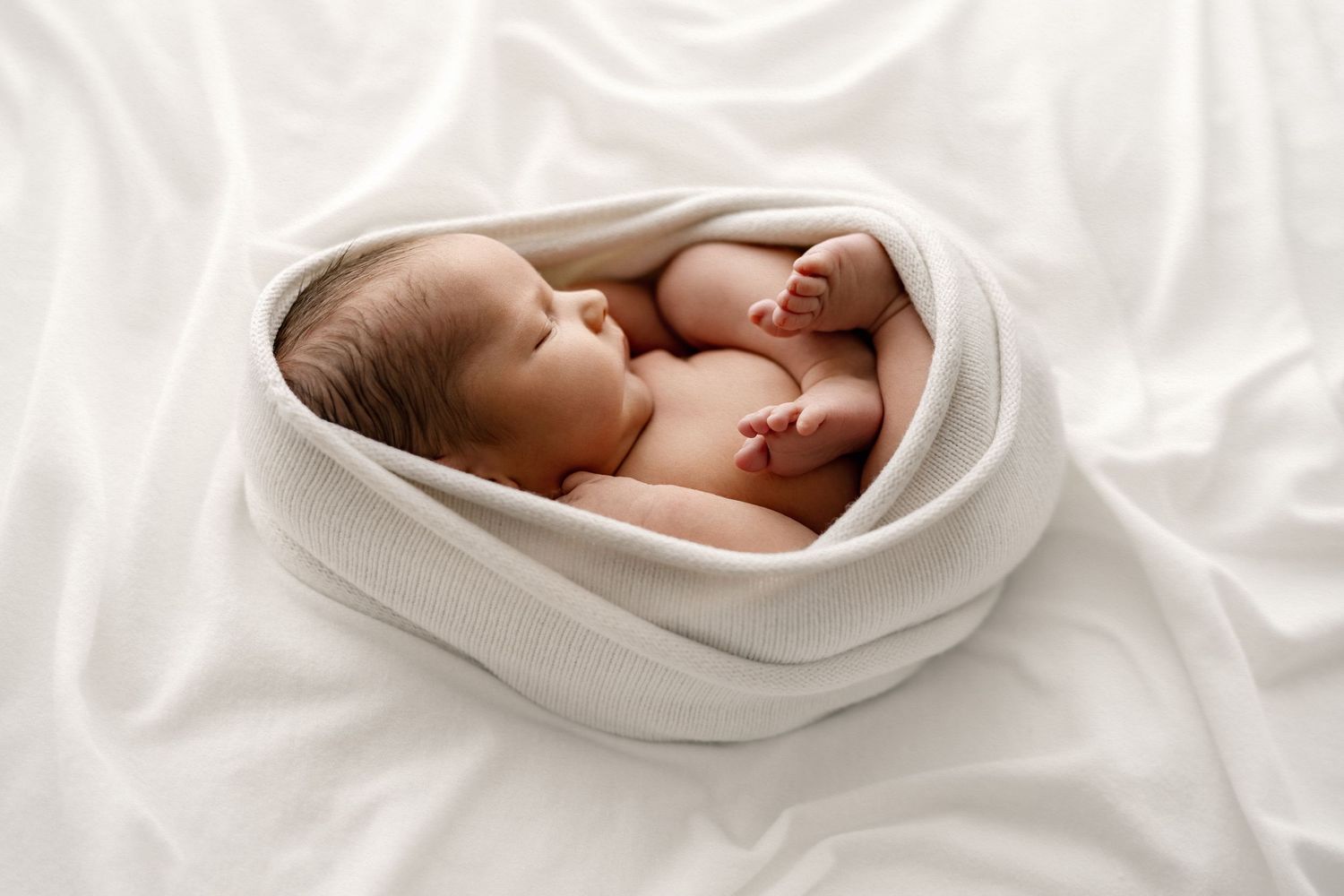 Close-up photo of tiny baby hand gently held in adult hands with white sleeve visible in soft lighting.