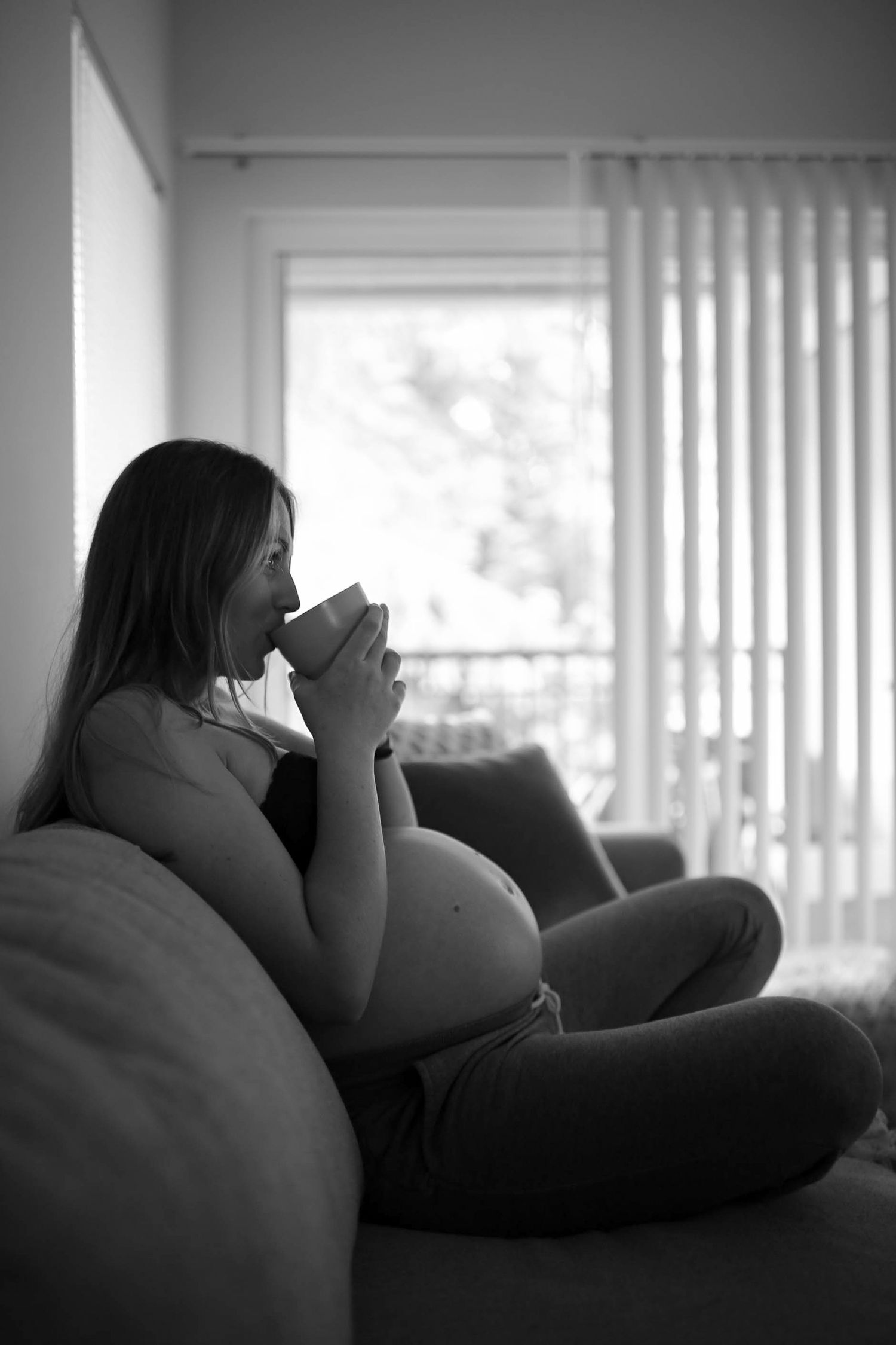Black and white silhouette of pregnant woman drinking from mug by window.