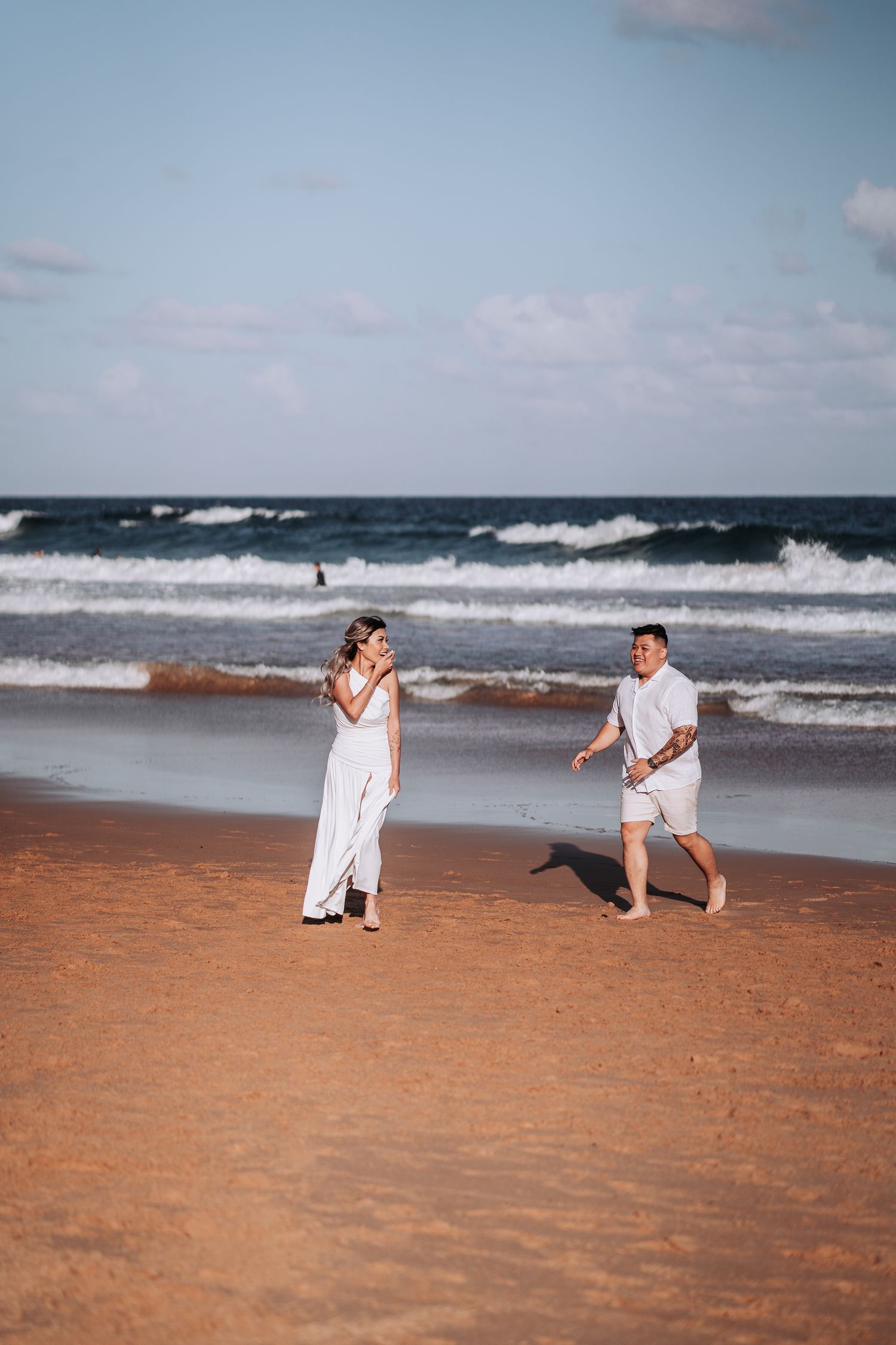 Two people walking along beach shoreline with crashing waves in background.