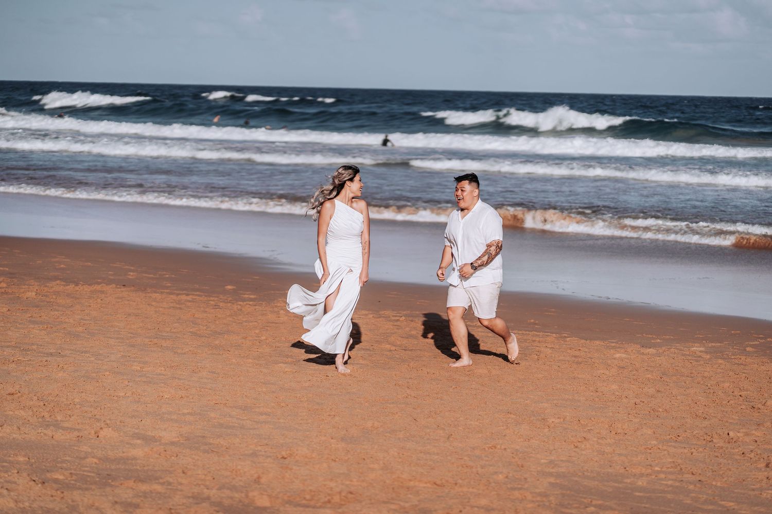 Two people in white outfits enjoying a beach walk with waves crashing behind them.