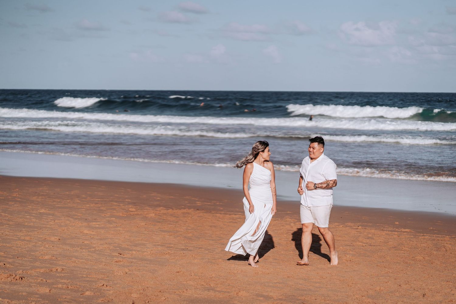 A couple in white outfits walks together along a sandy beach with ocean waves in the background.