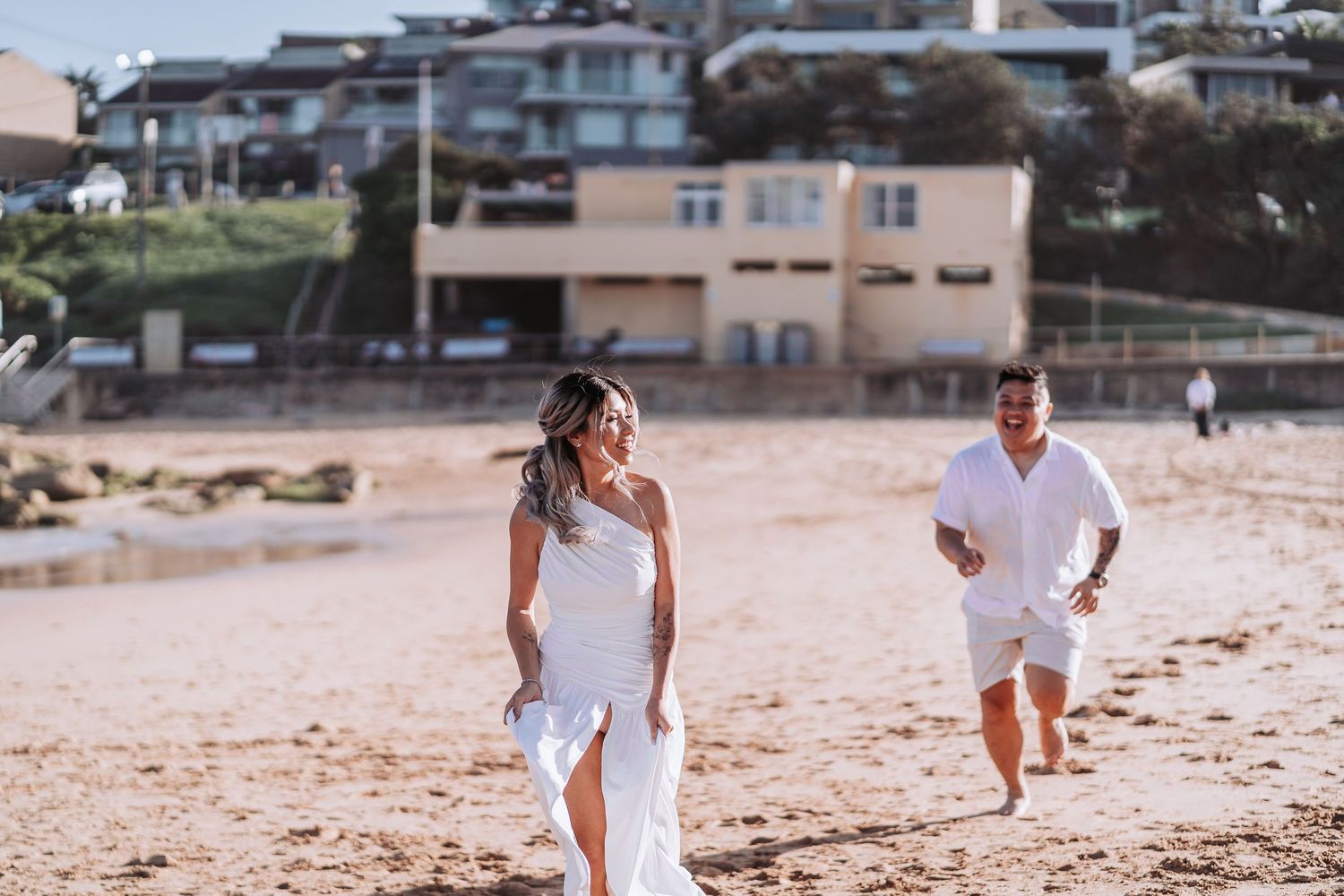 Couple running playfully on sandy beach with coastal houses visible in background.