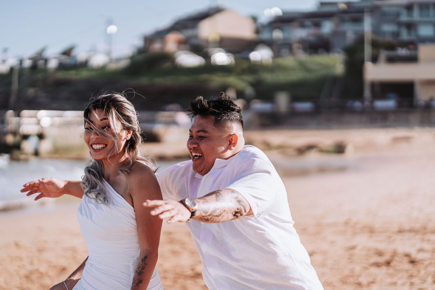 Two people in matching white outfits laugh and play together on a sunny beach.
