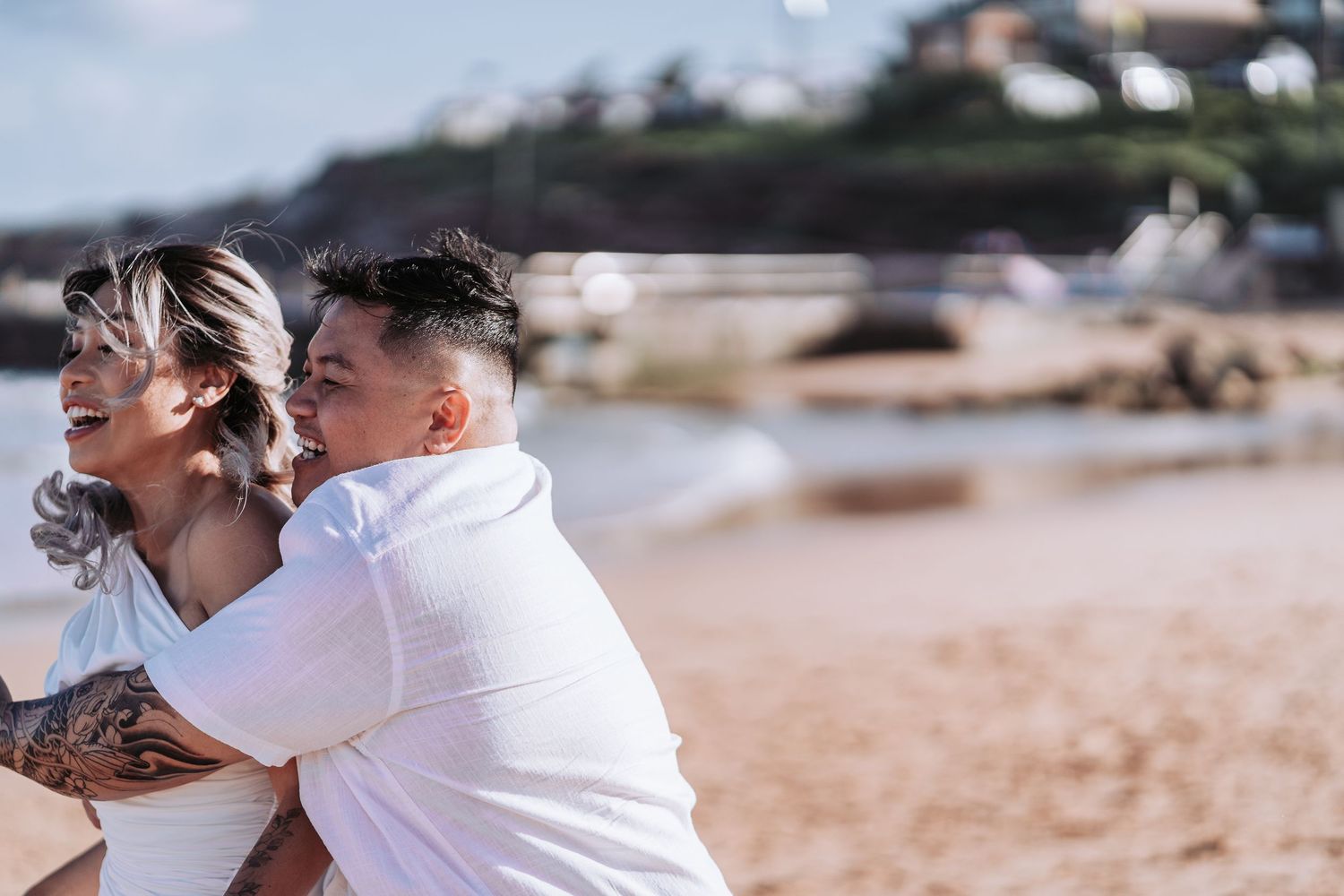 Joyful couple in white embracing and laughing on a beach with coastal homes in the background.