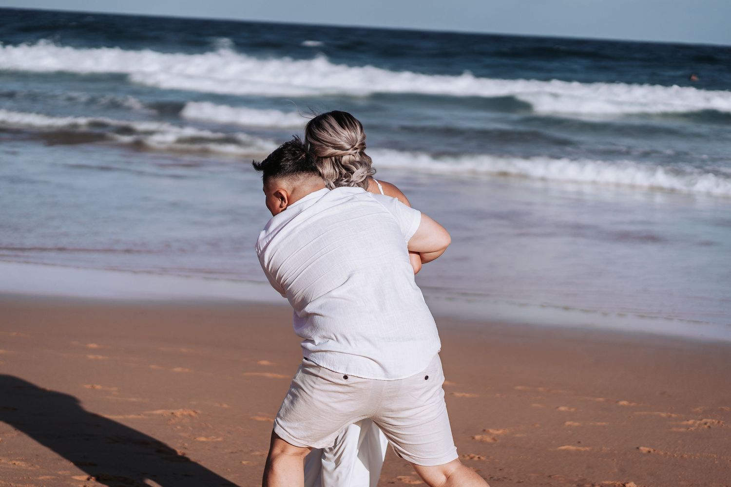 Couple in white clothing share an embrace on a sandy beach with crashing waves.