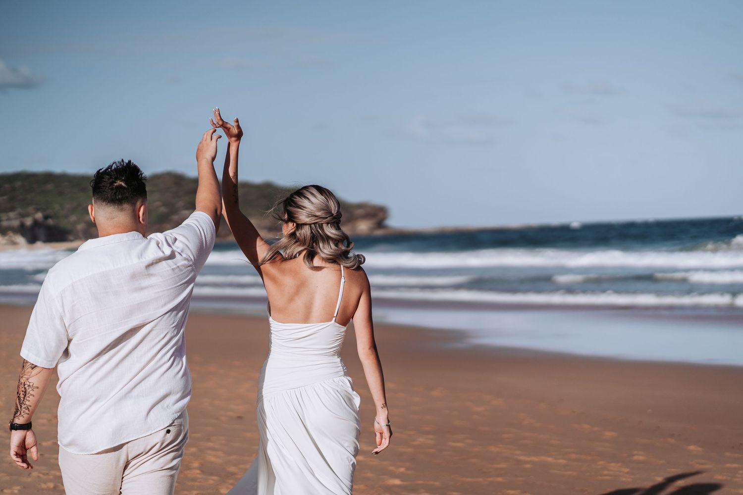 Couple in white outfits holding hands while walking on beach with waves and houses in background.