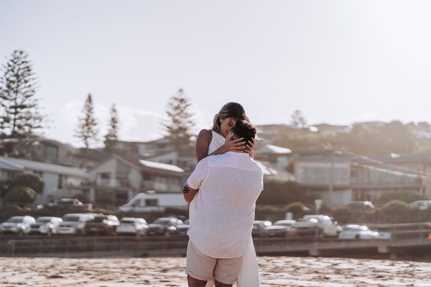 Romantic beach embrace with coastal homes and pine trees in the background.