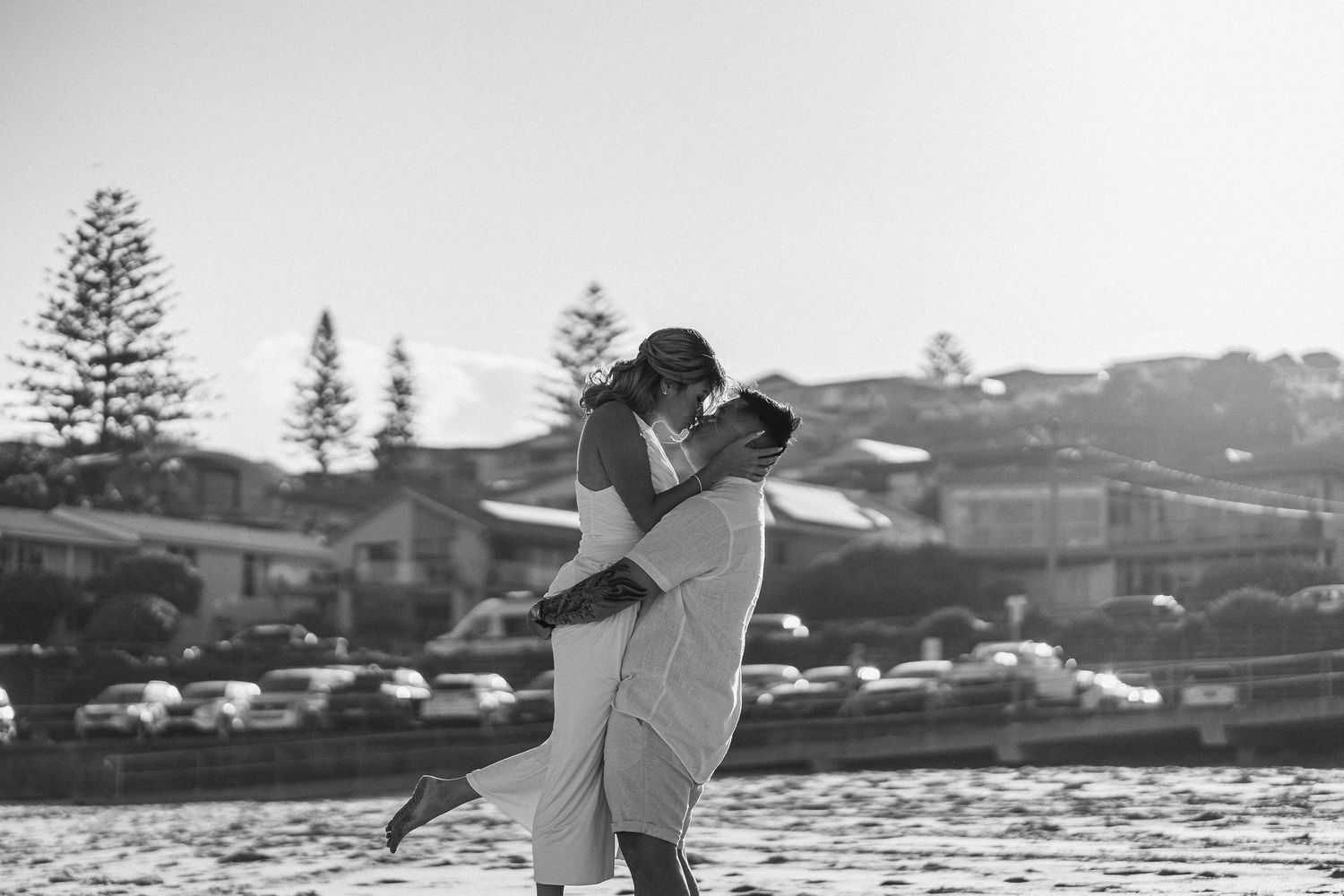 Black and white photo of romantic beach moment with coastal scenery in background.