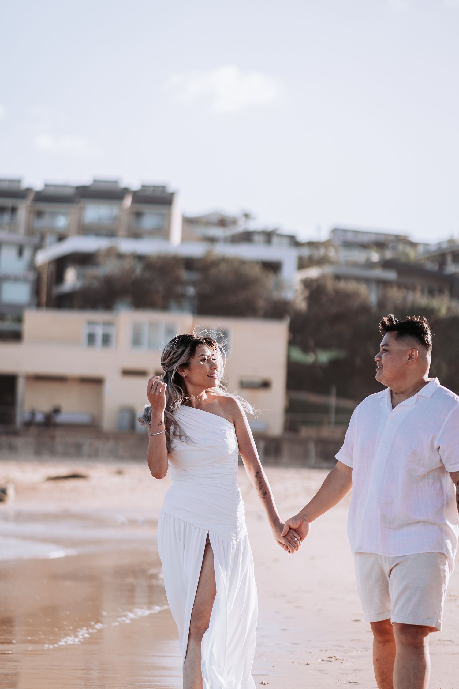 Couple in white attire holding hands on beach with seaside houses visible on hillside.
