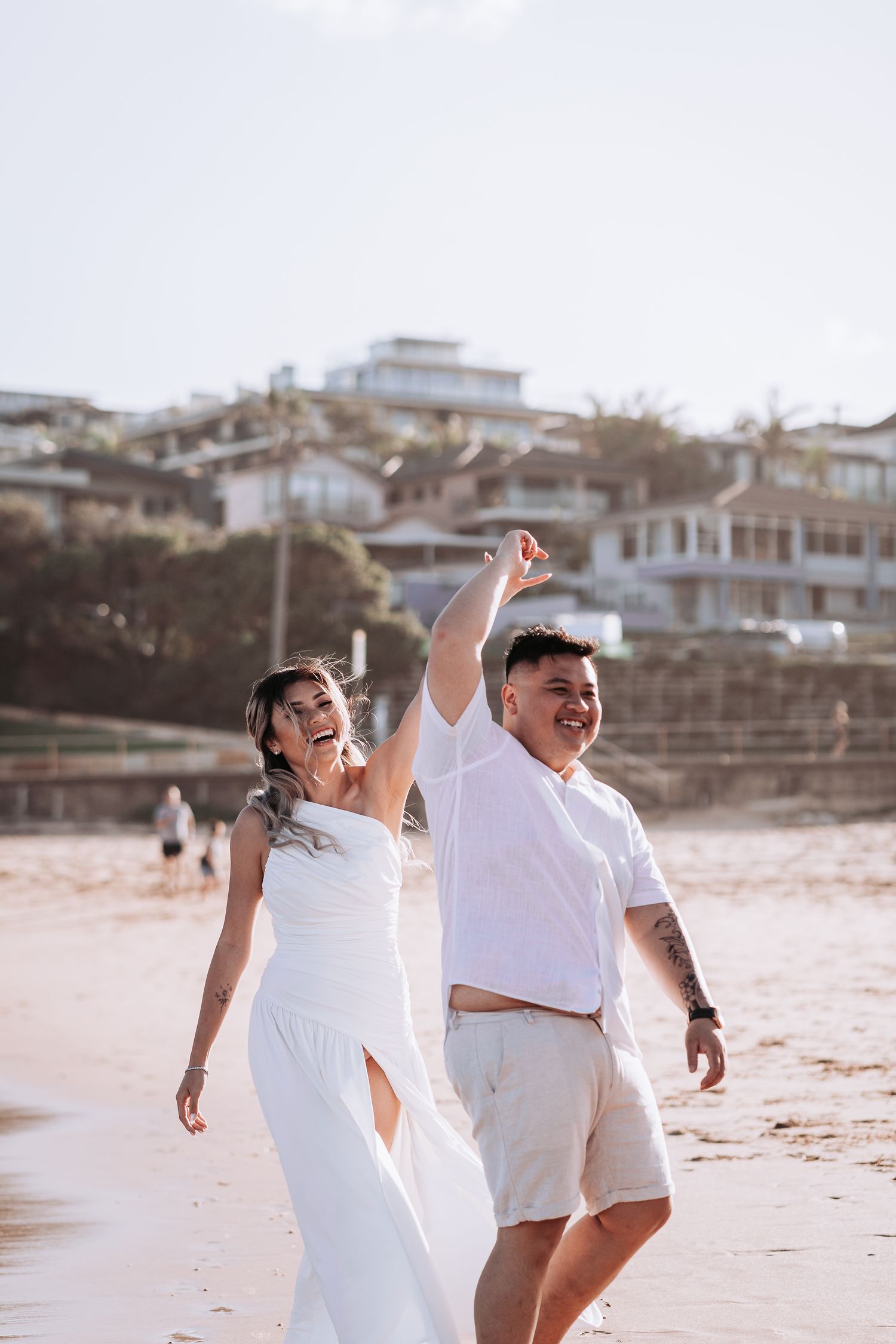 Dancing couple in white outfits enjoying a playful moment on the beach.
