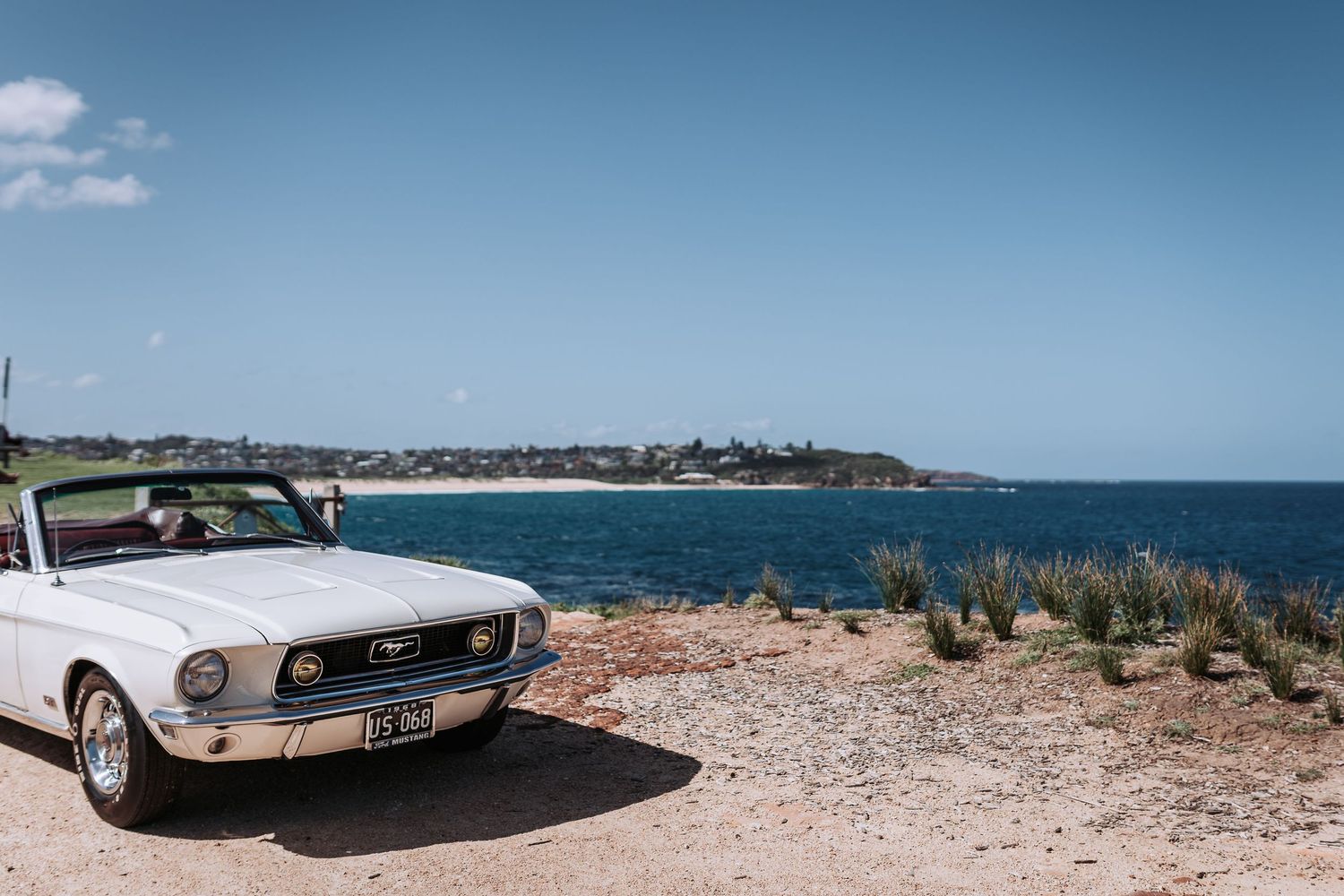 A vintage white Ford Mustang convertible parked on a rocky coastal overlook with blue ocean waters.
