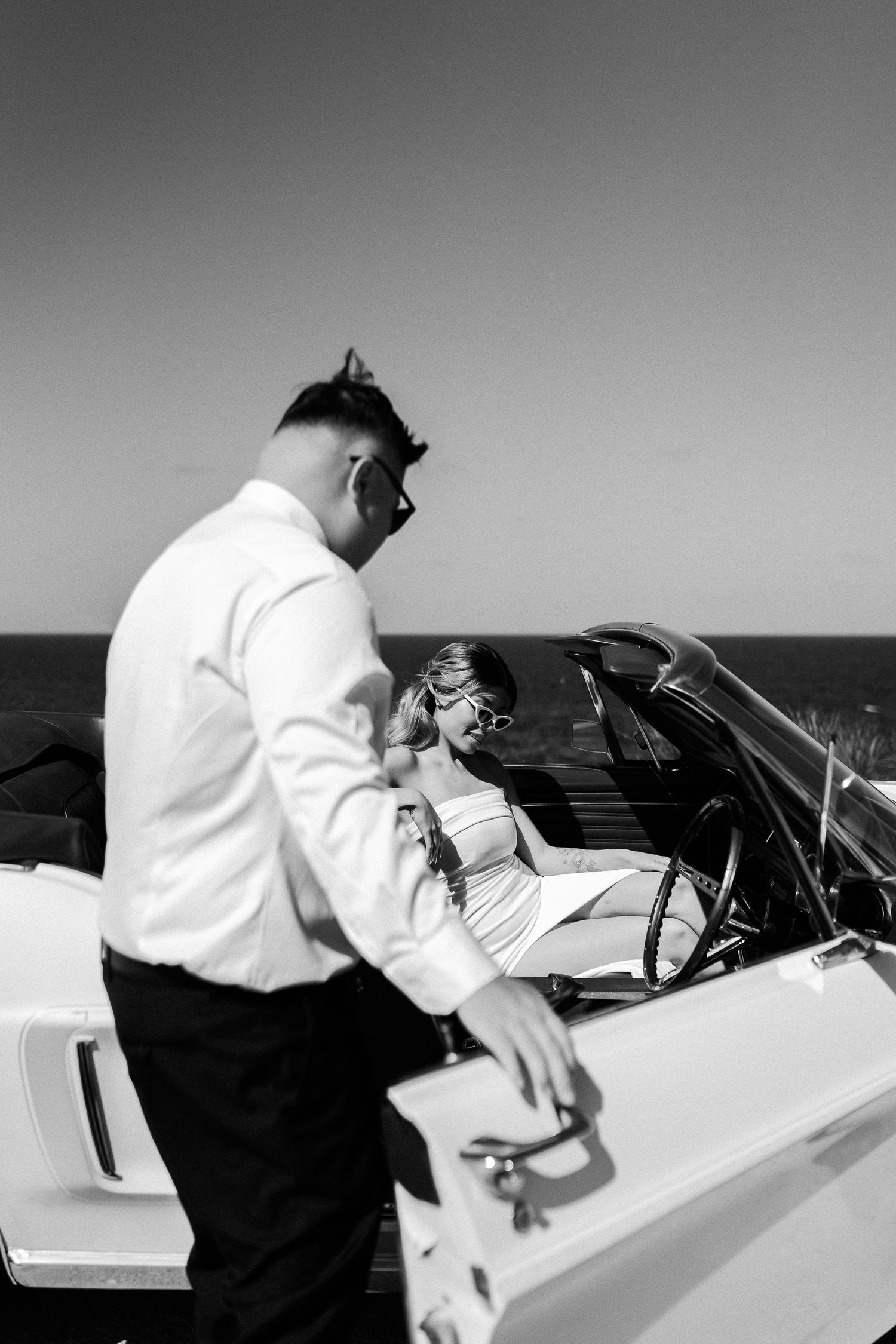 Black and white photo of a couple seated in a vintage convertible car by the waterfront.