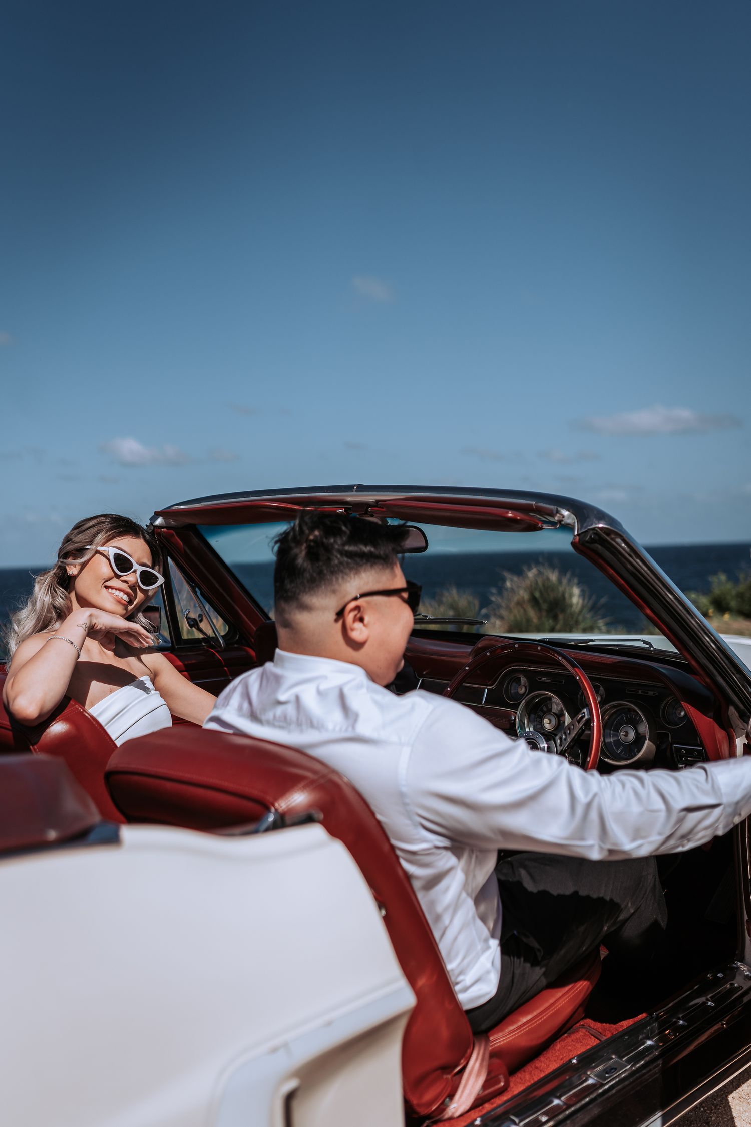 A couple enjoying a sunny day in a classic white convertible with red interior by the ocean.
