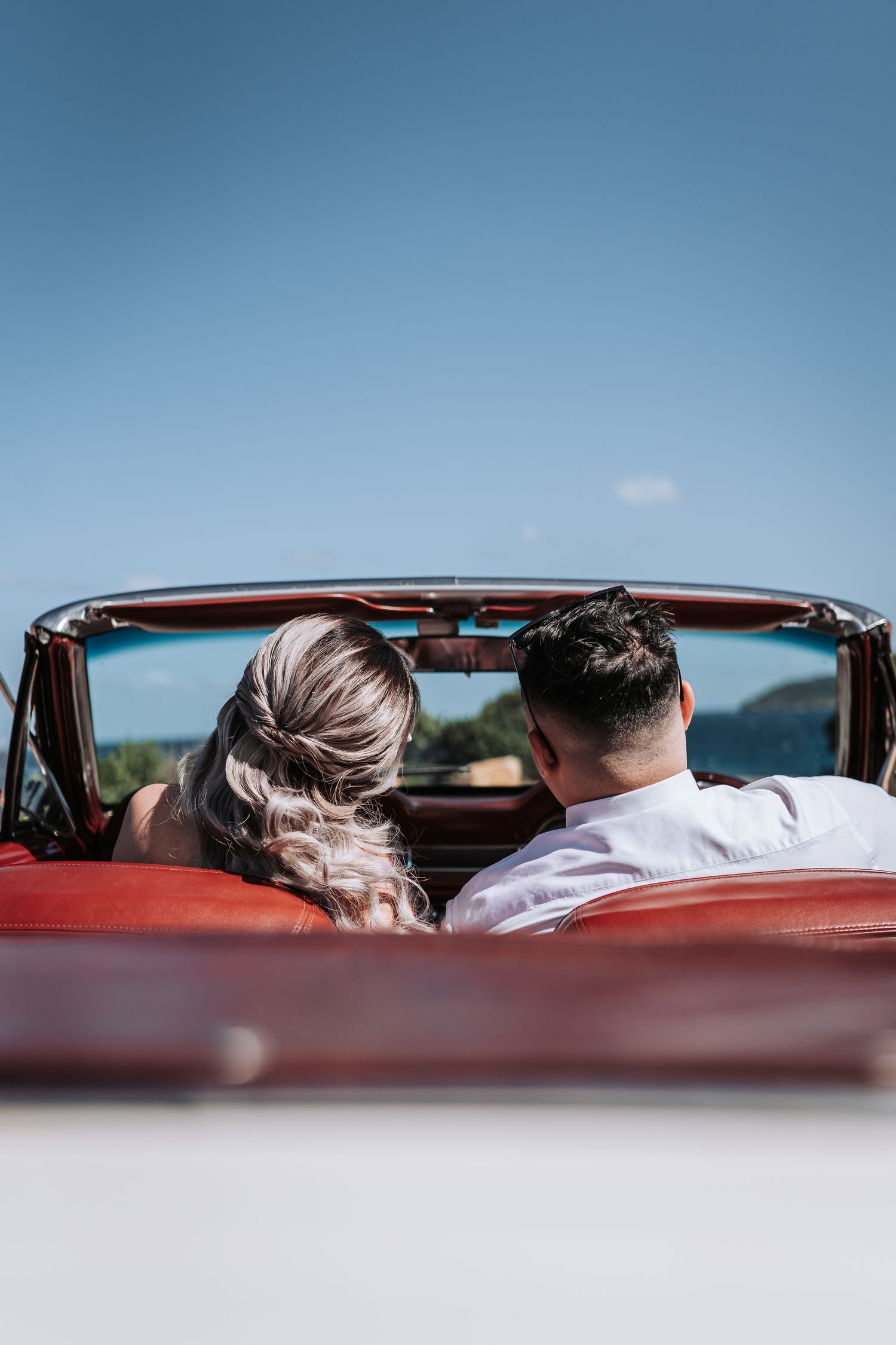 Back view of a couple seated in a white classic car with red leather interior against blue sky.