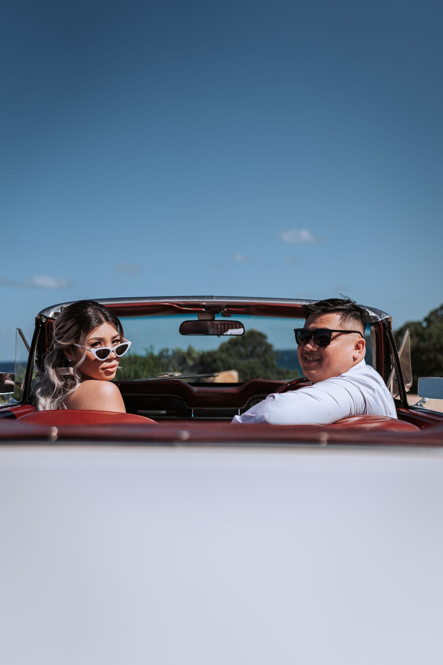 A stylish couple in sunglasses seated in a vintage white convertible with red interior.