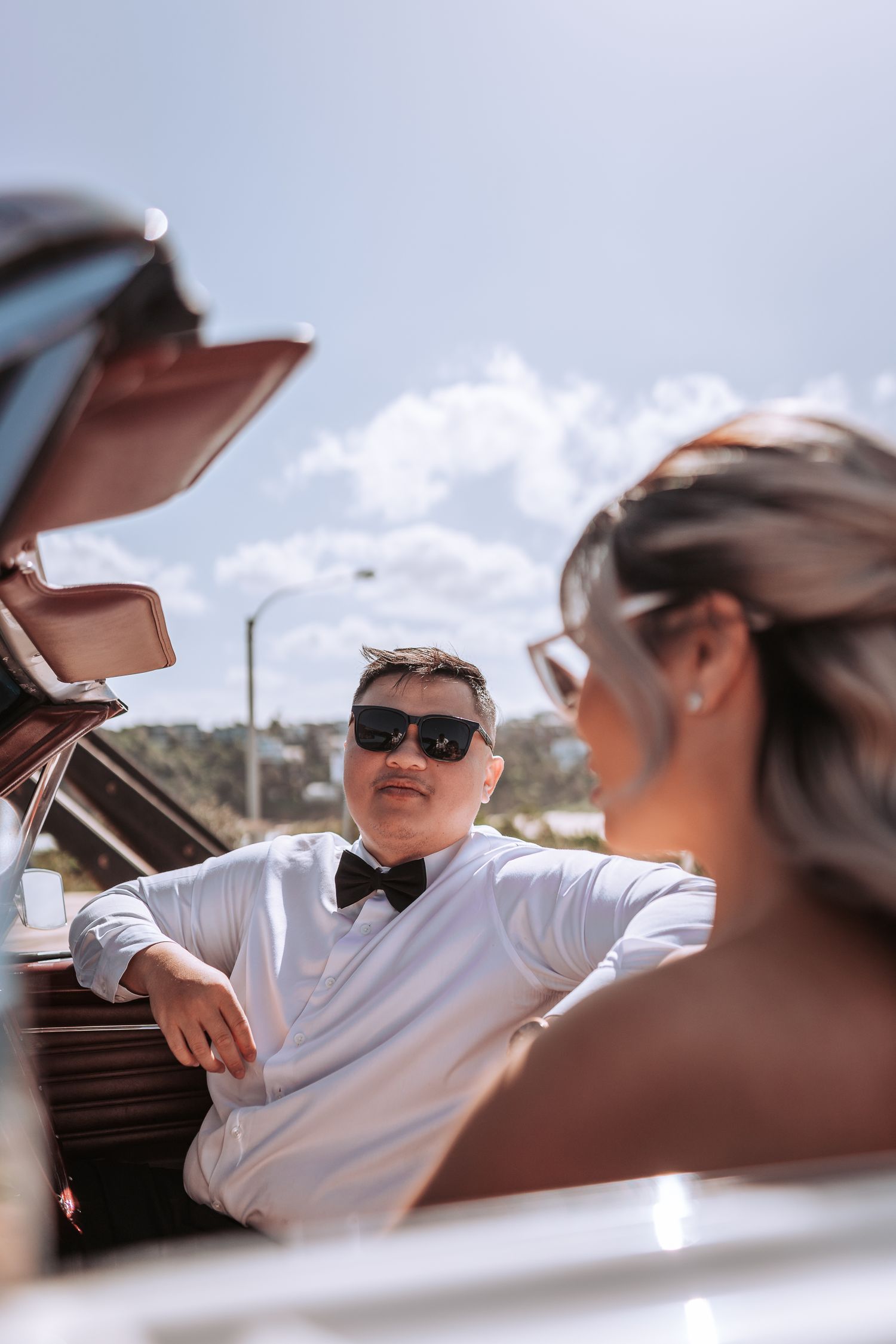 Elegant couple in formal attire posed in vintage convertible during sunny outdoor photoshoot.
