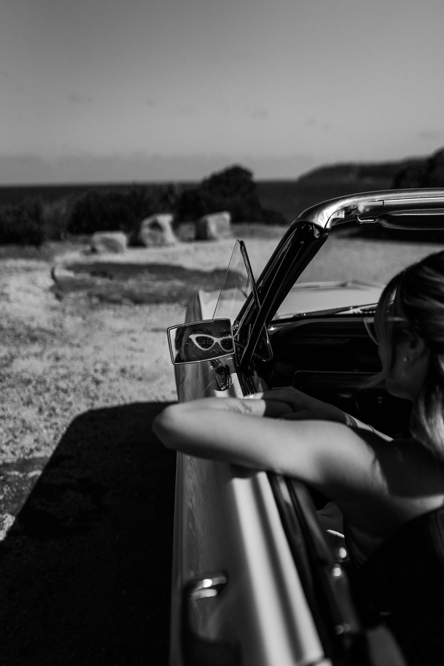 Artistic black and white shot of vintage car details with sunglasses and coastal backdrop.