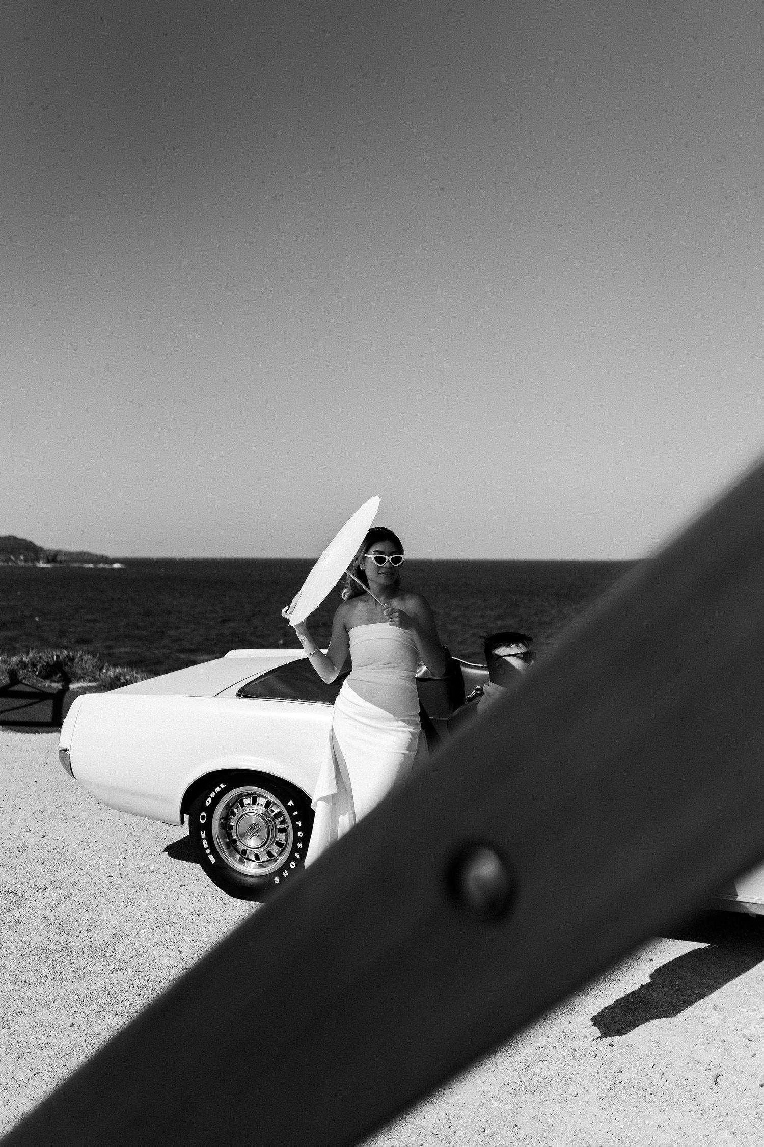 A black and white portrait with a classic convertible car against an ocean backdrop.