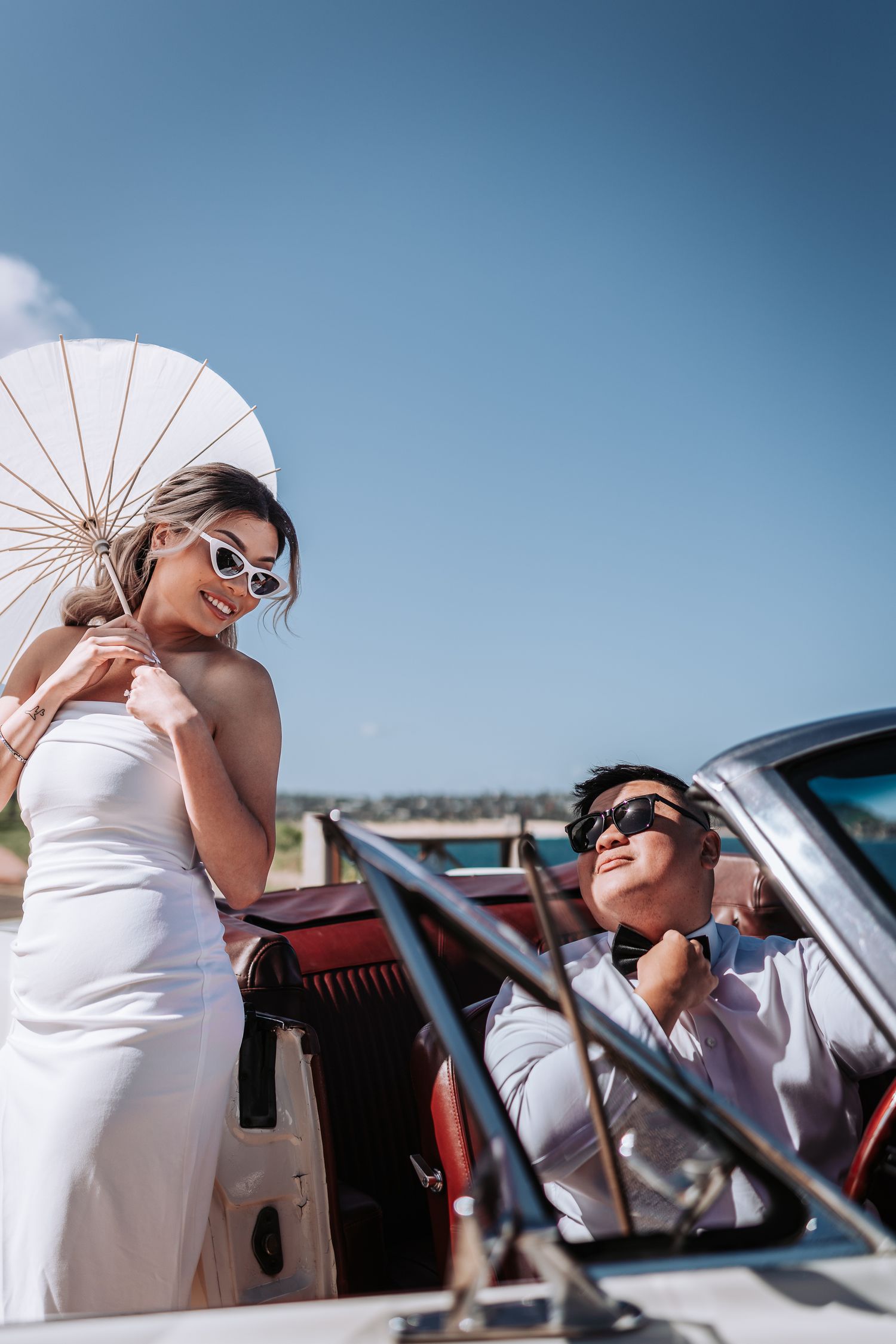 A stylish couple poses with a white parasol by a classic car on a coastal road.
