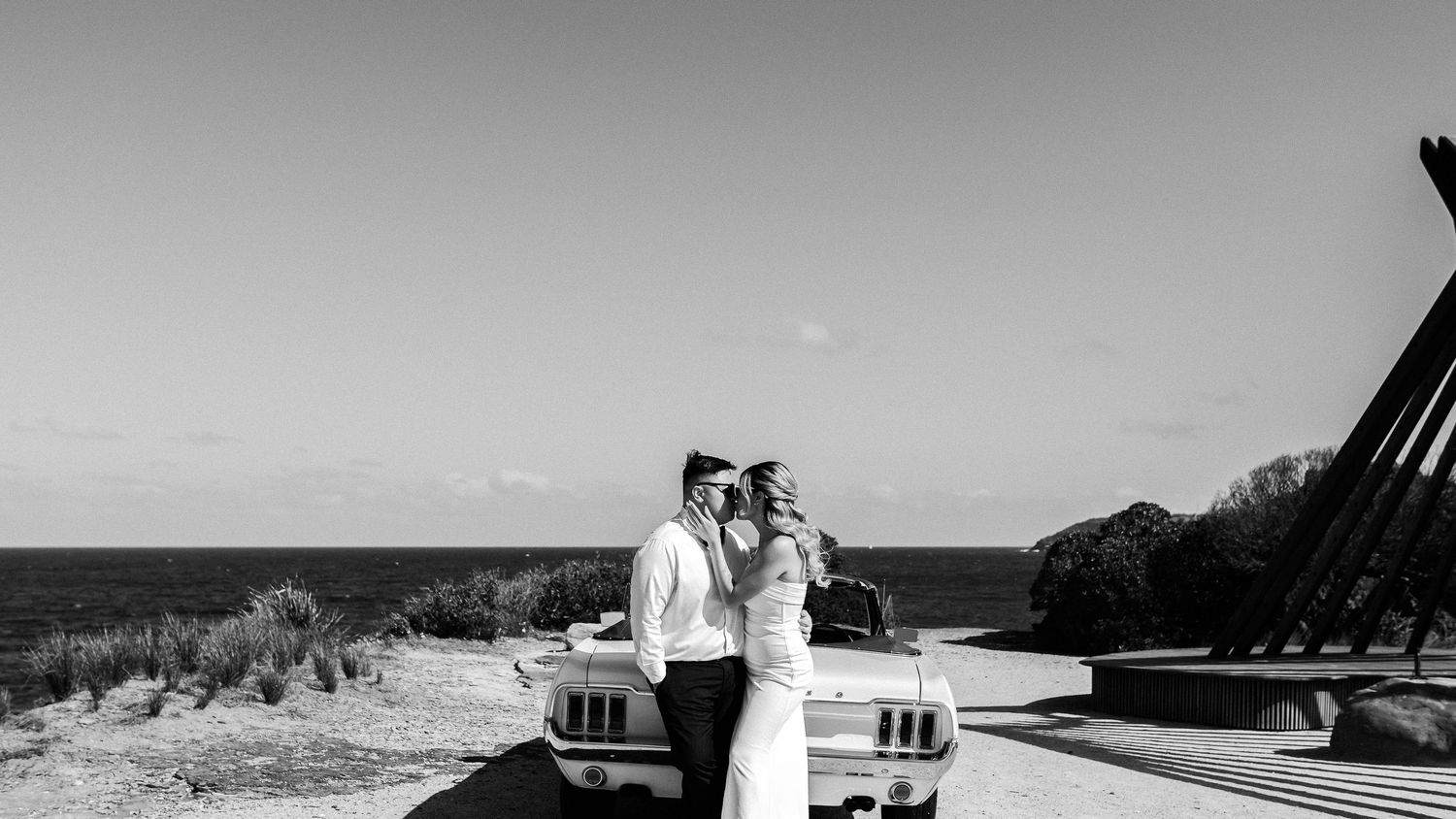 A romantic black and white photo of newlyweds posing with a classic car by the ocean.
