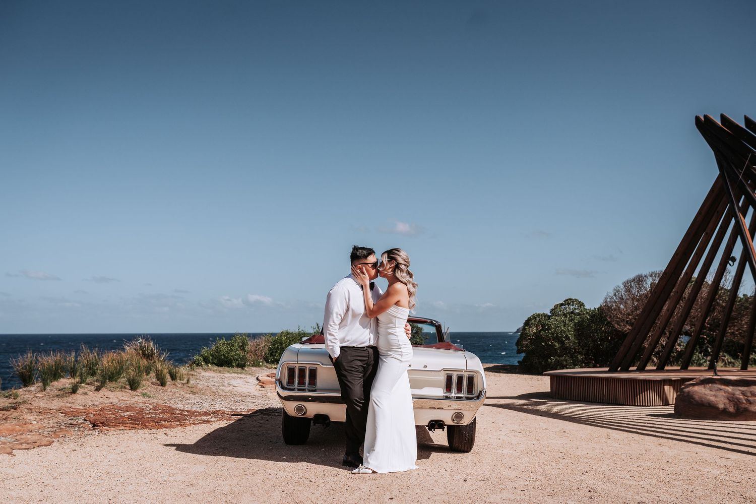 Newlyweds share a romantic moment next to a vintage car with the ocean in the background.