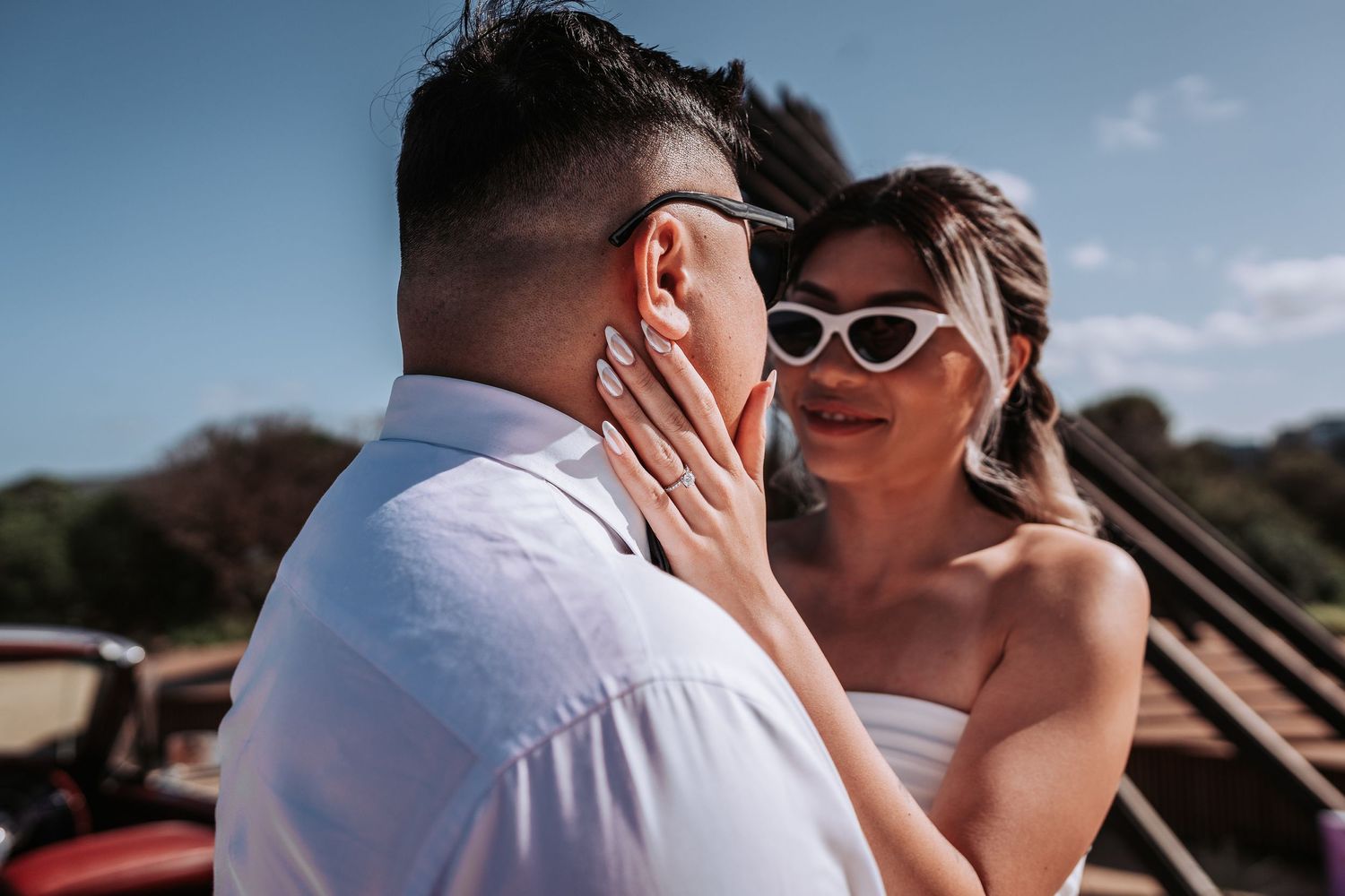 A tender moment captured between a couple in wedding attire with sunglasses near the coast.