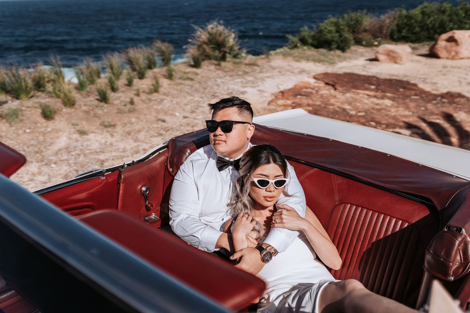 Stylish wedding couple wearing sunglasses while sitting in a red leather vintage convertible.