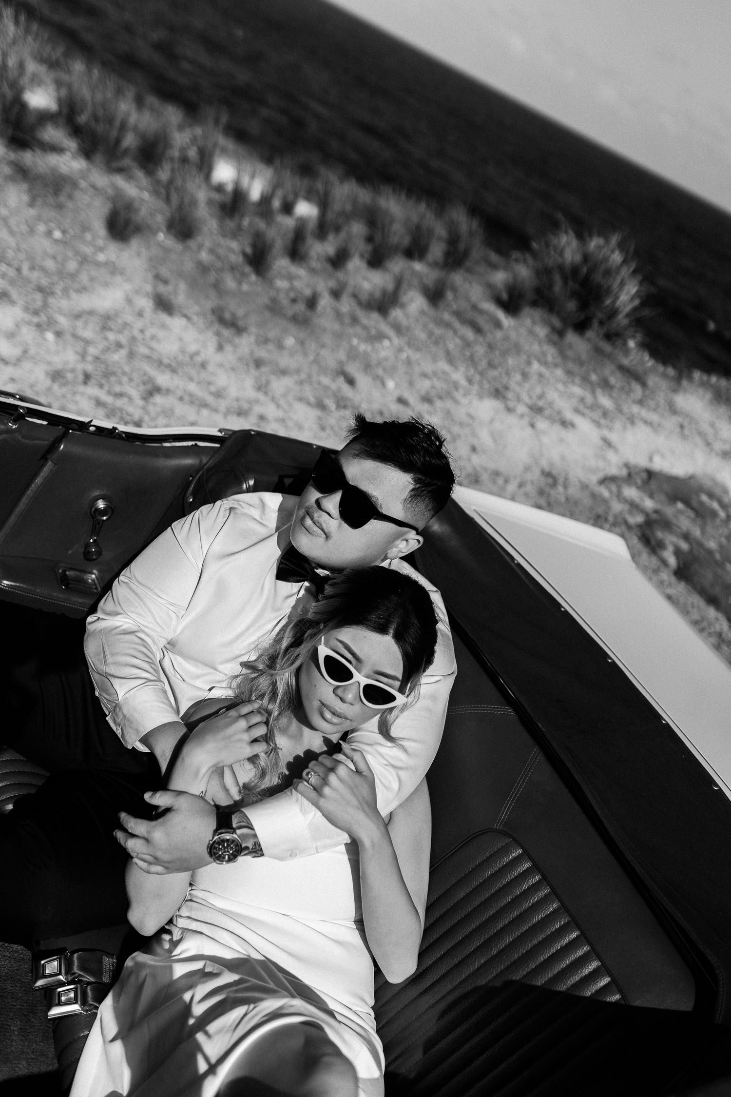 Black and white photo of couple lounging in classic convertible car with retro style sunglasses.