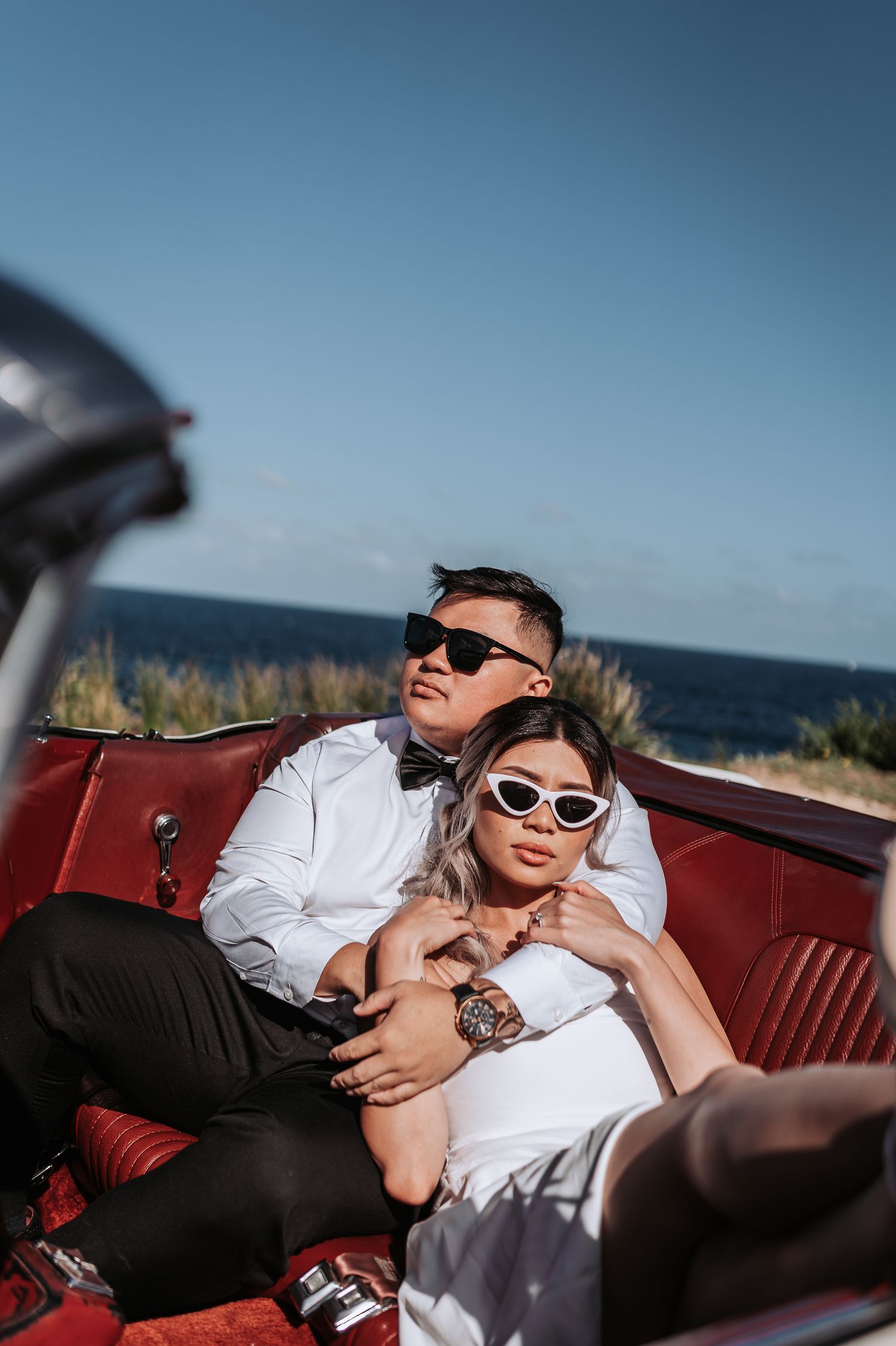 Couple in formal attire relaxing in vintage red convertible car by the ocean wearing sunglasses.