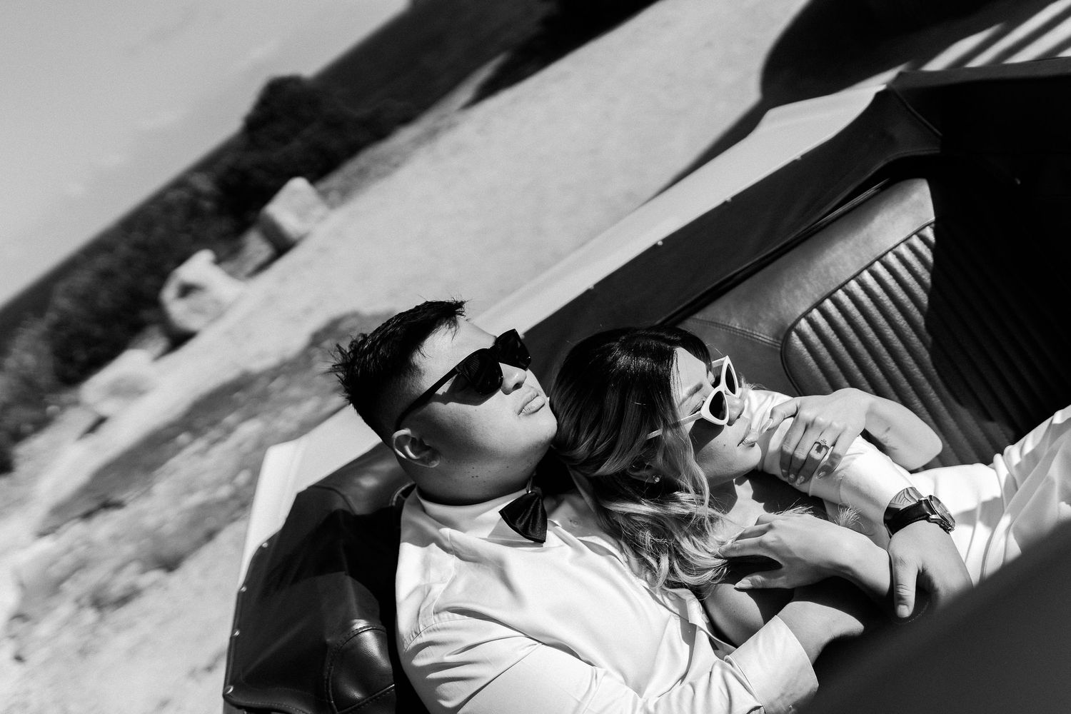 Dramatic black and white photo of couple embracing in vintage convertible with scenic coastal backdrop.