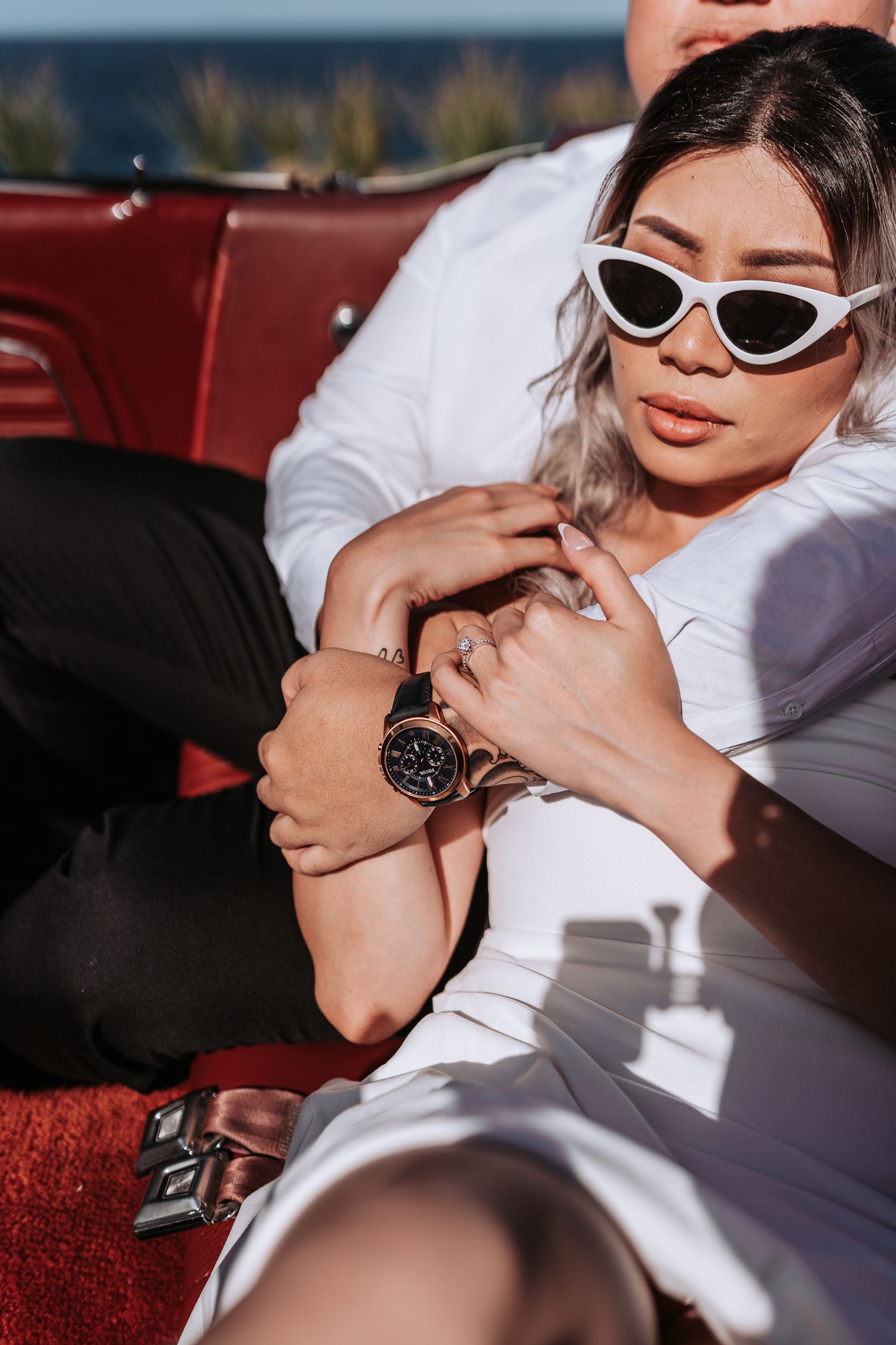 Close up of couple in white outfits cuddling in red vintage car wearing stylish sunglasses.