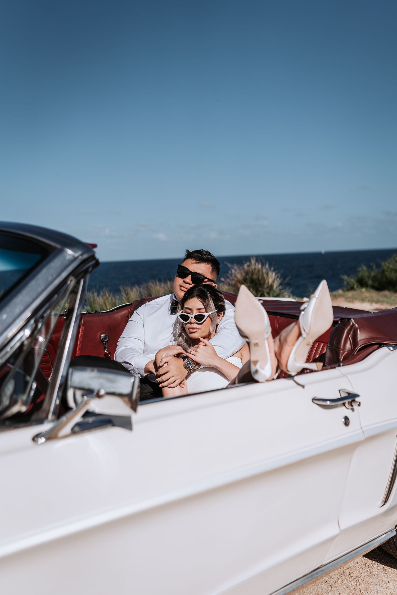 Couple in formal wear posing romantically in white vintage convertible by oceanside.