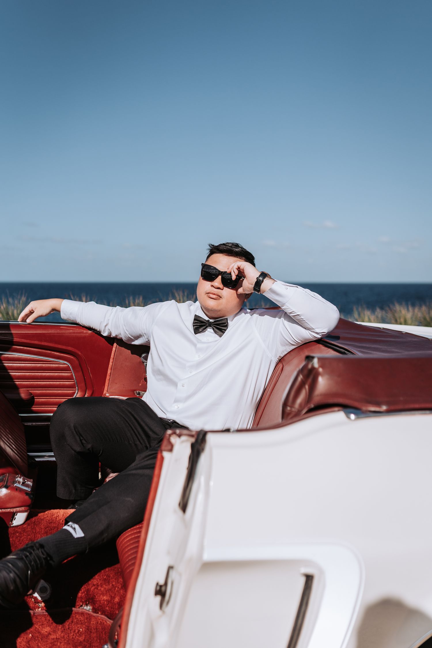 Man in white shirt and bowtie posing in classic red convertible against blue ocean backdrop.