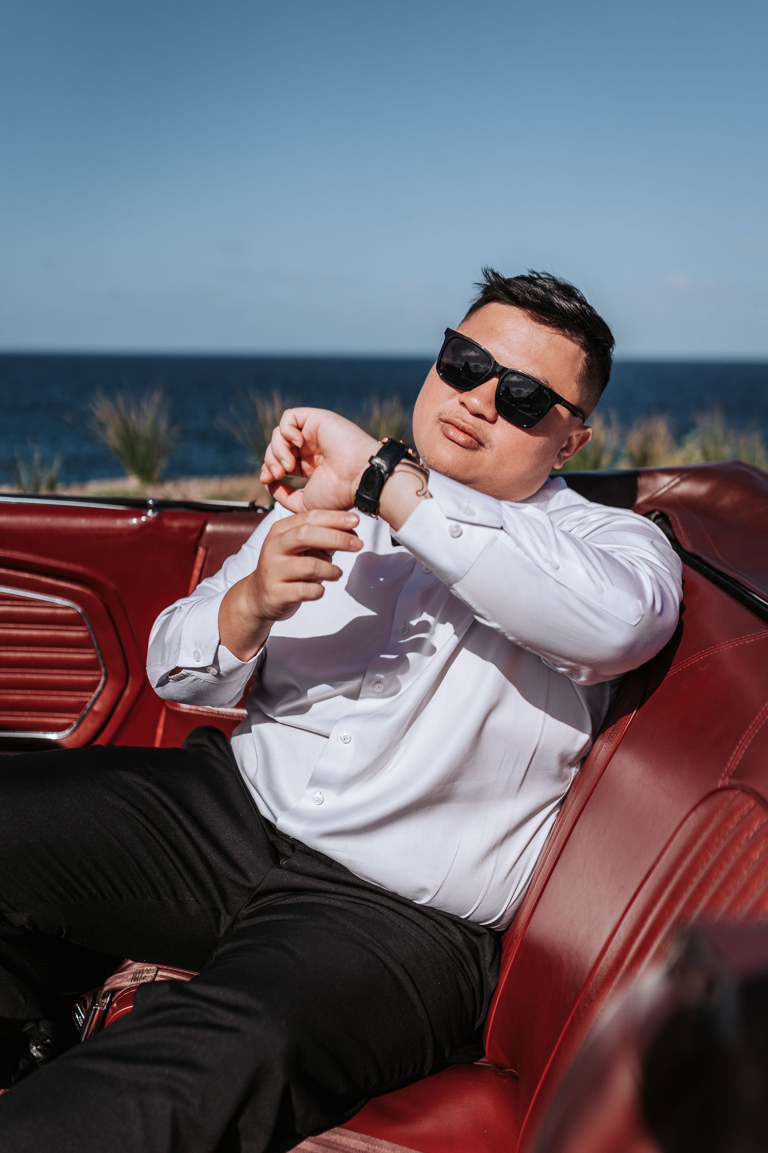 Person in white shirt relaxing in red vintage car seat near ocean.