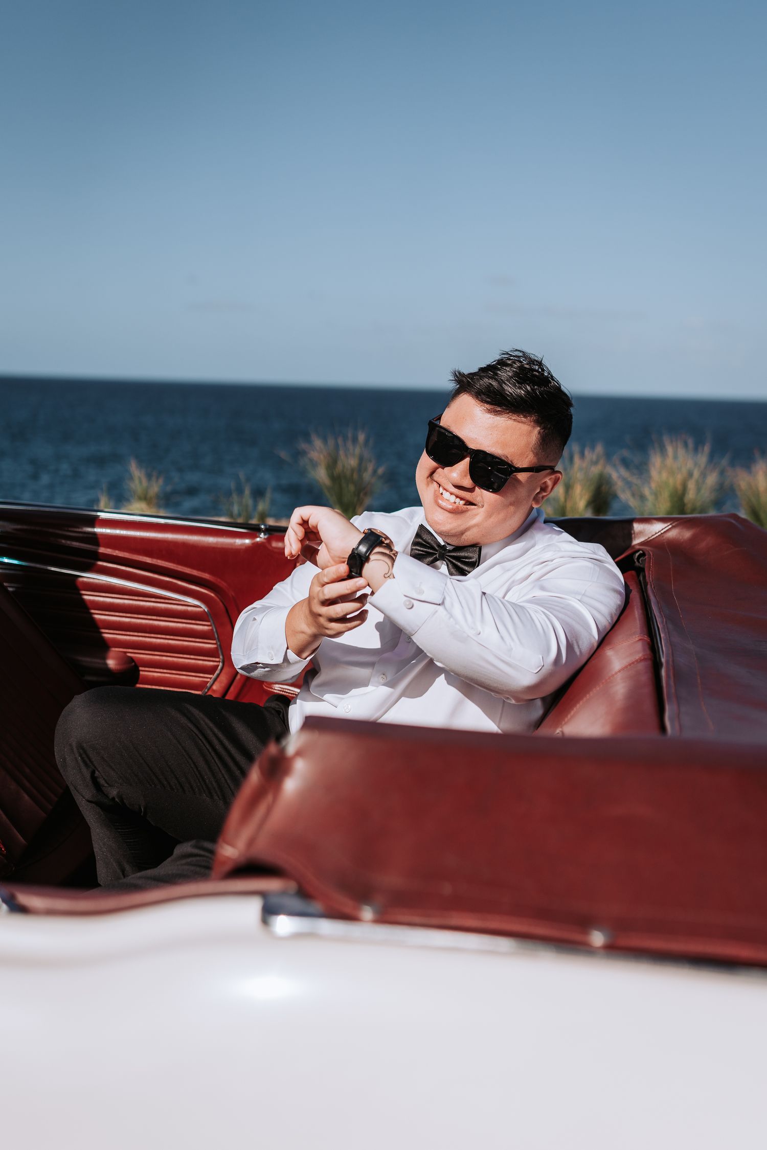 Man in formal wear checking watch while seated in vintage convertible by ocean.