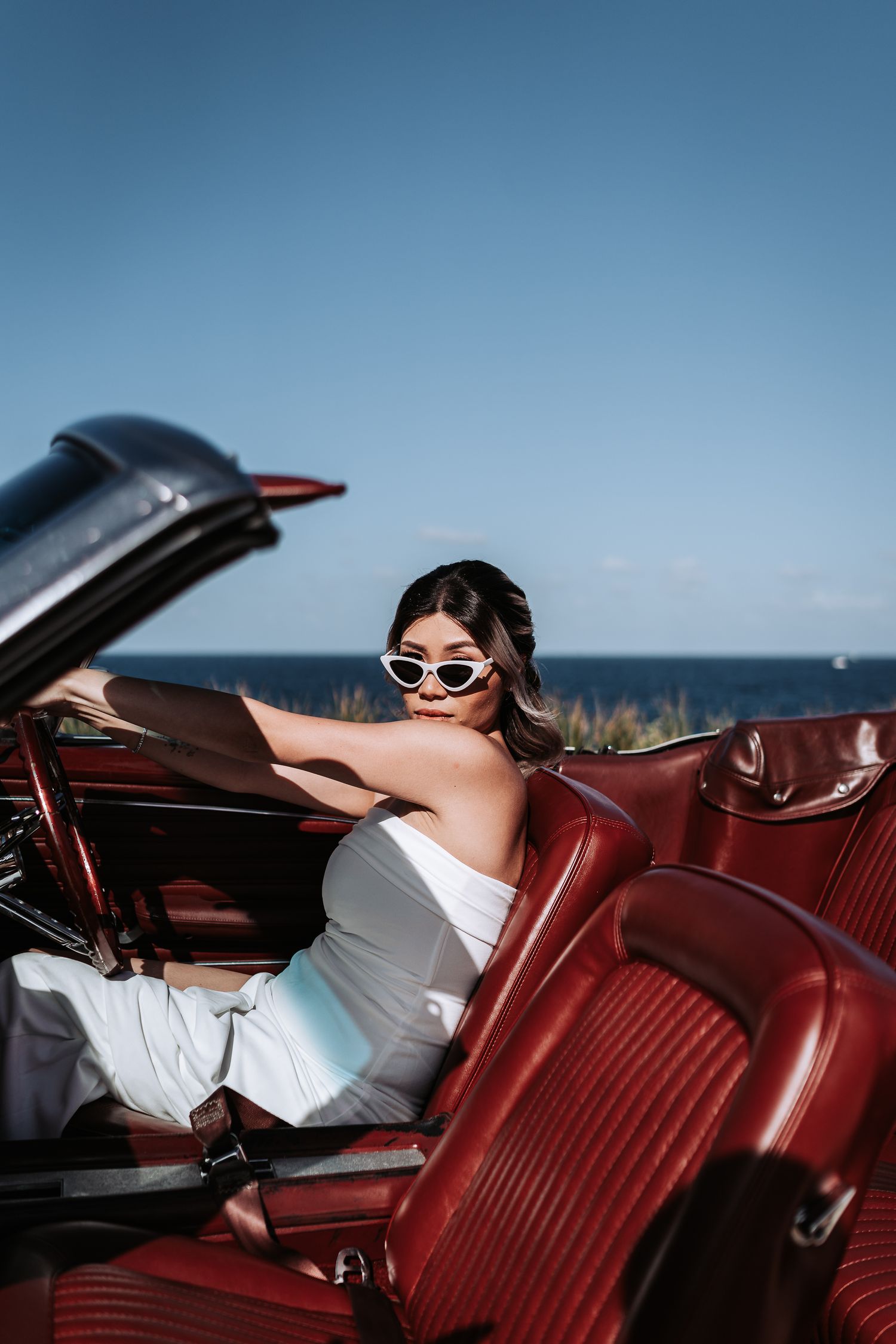 Woman in sunglasses posing in red vintage convertible by seaside.