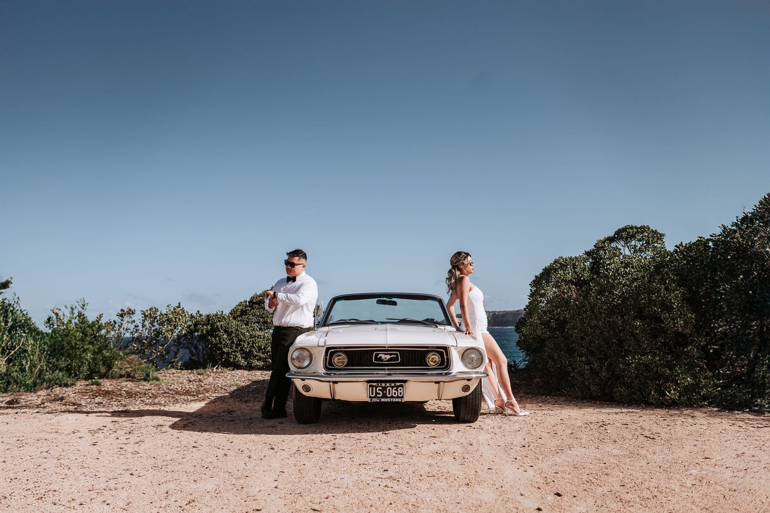 Couple poses with vintage white Mustang convertible on scenic coastal cliff.