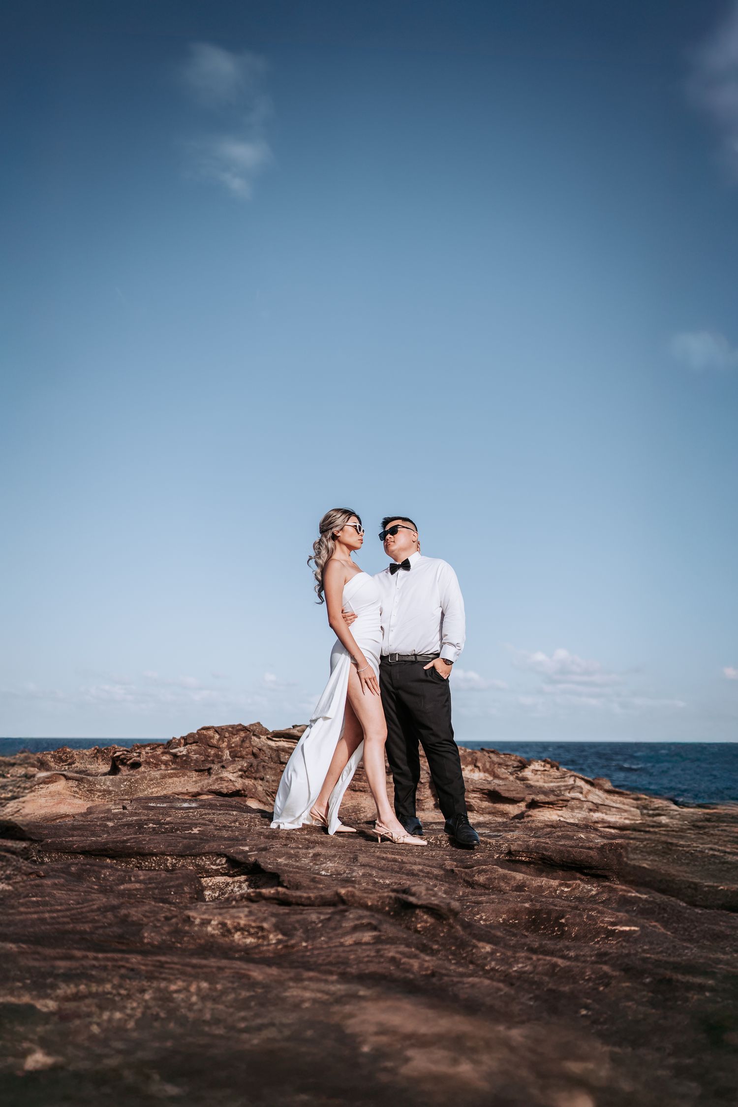 Elegant couple poses on rocky oceanside cliff under blue sky.