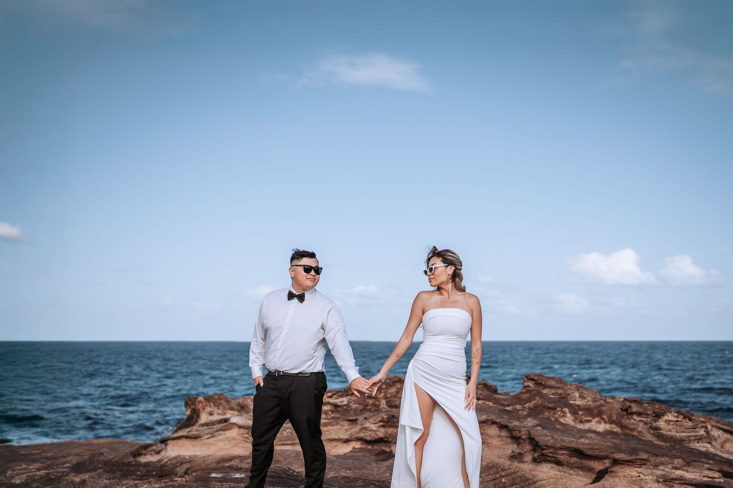 Couple holding hands on rocky oceanfront with blue horizon background.