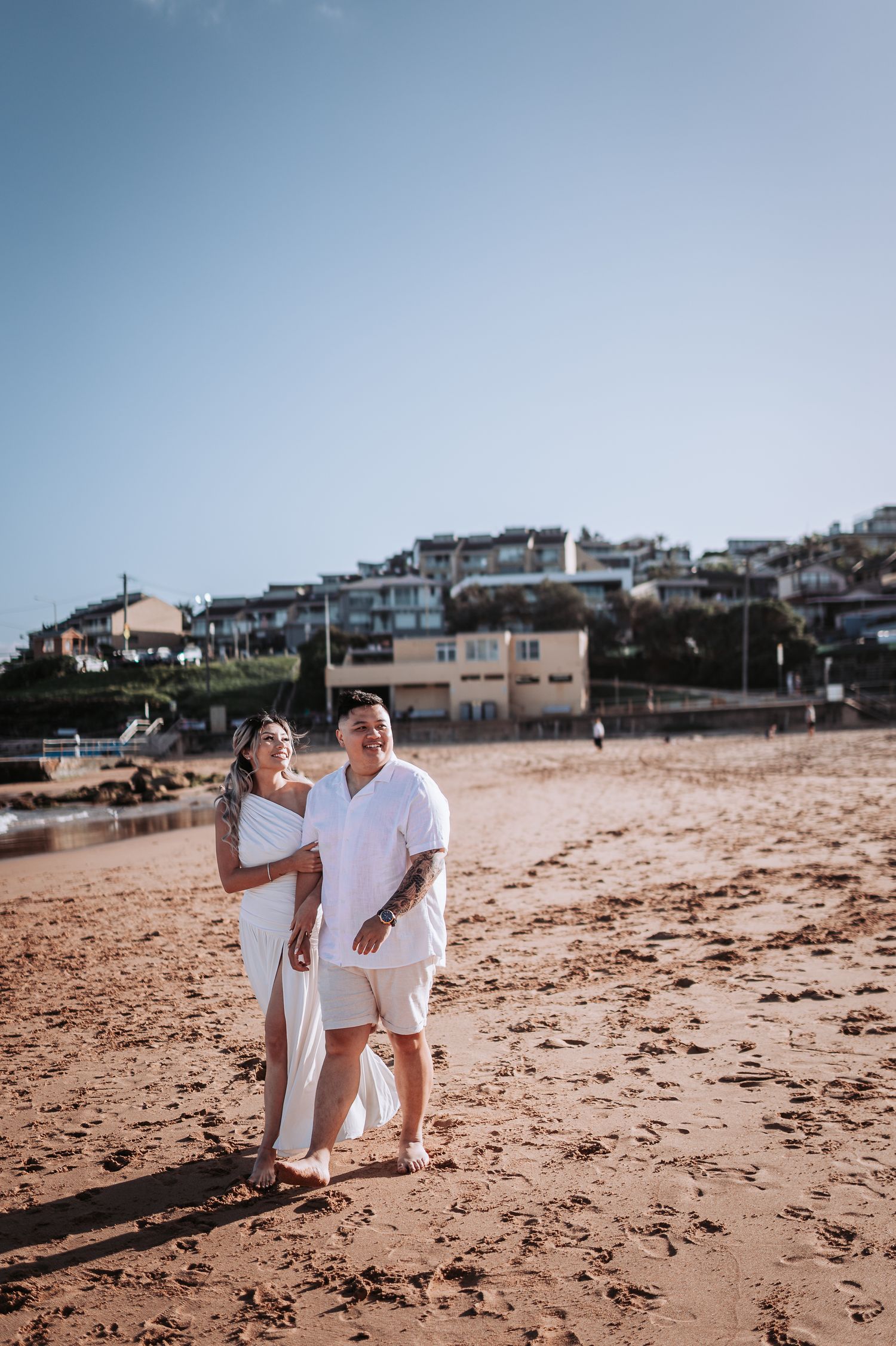 Couple in white attire standing together on sandy beach with coastal homes in background.