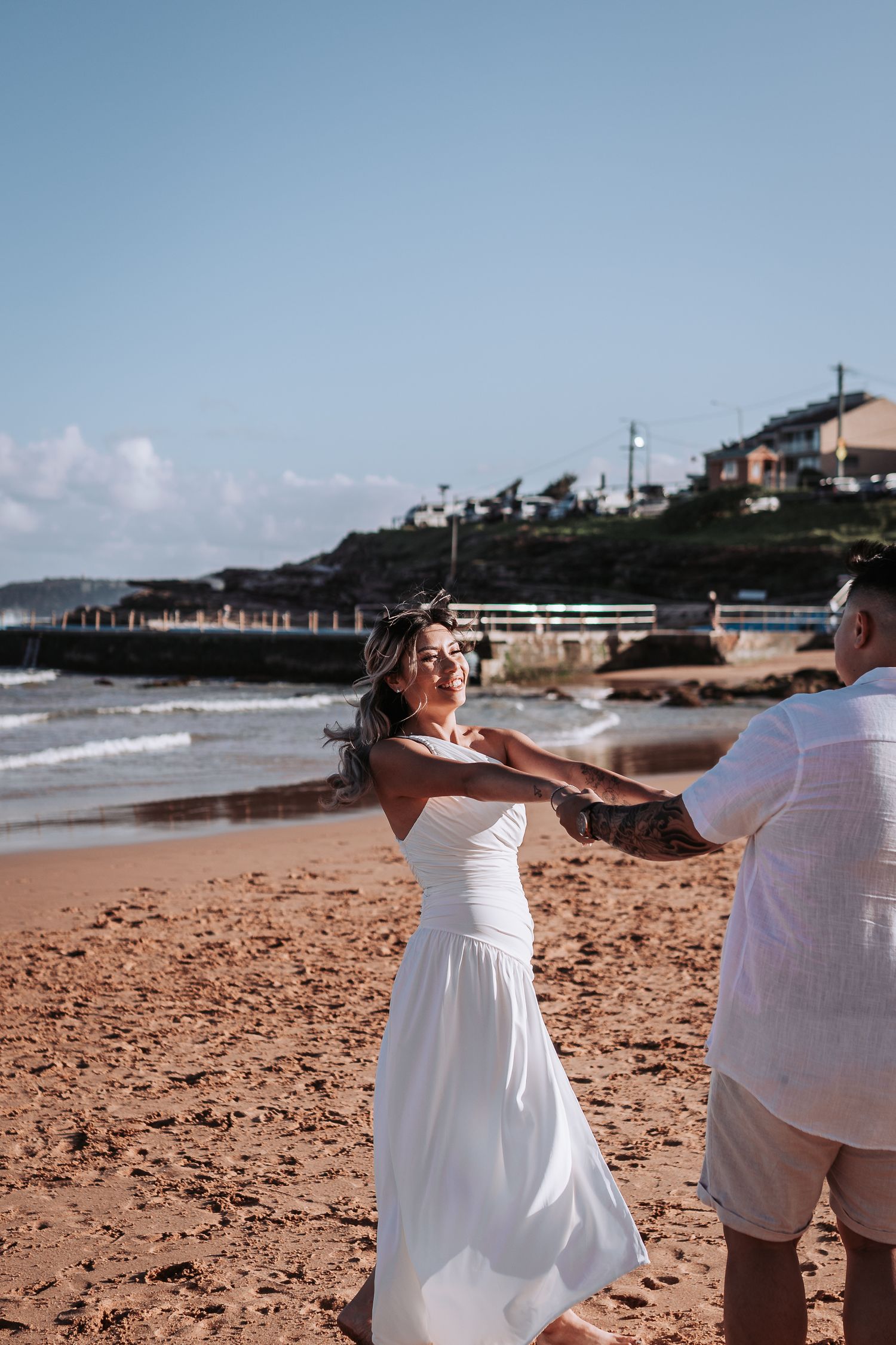 Two figures dancing together on beach with white clothing and seaside homes visible.