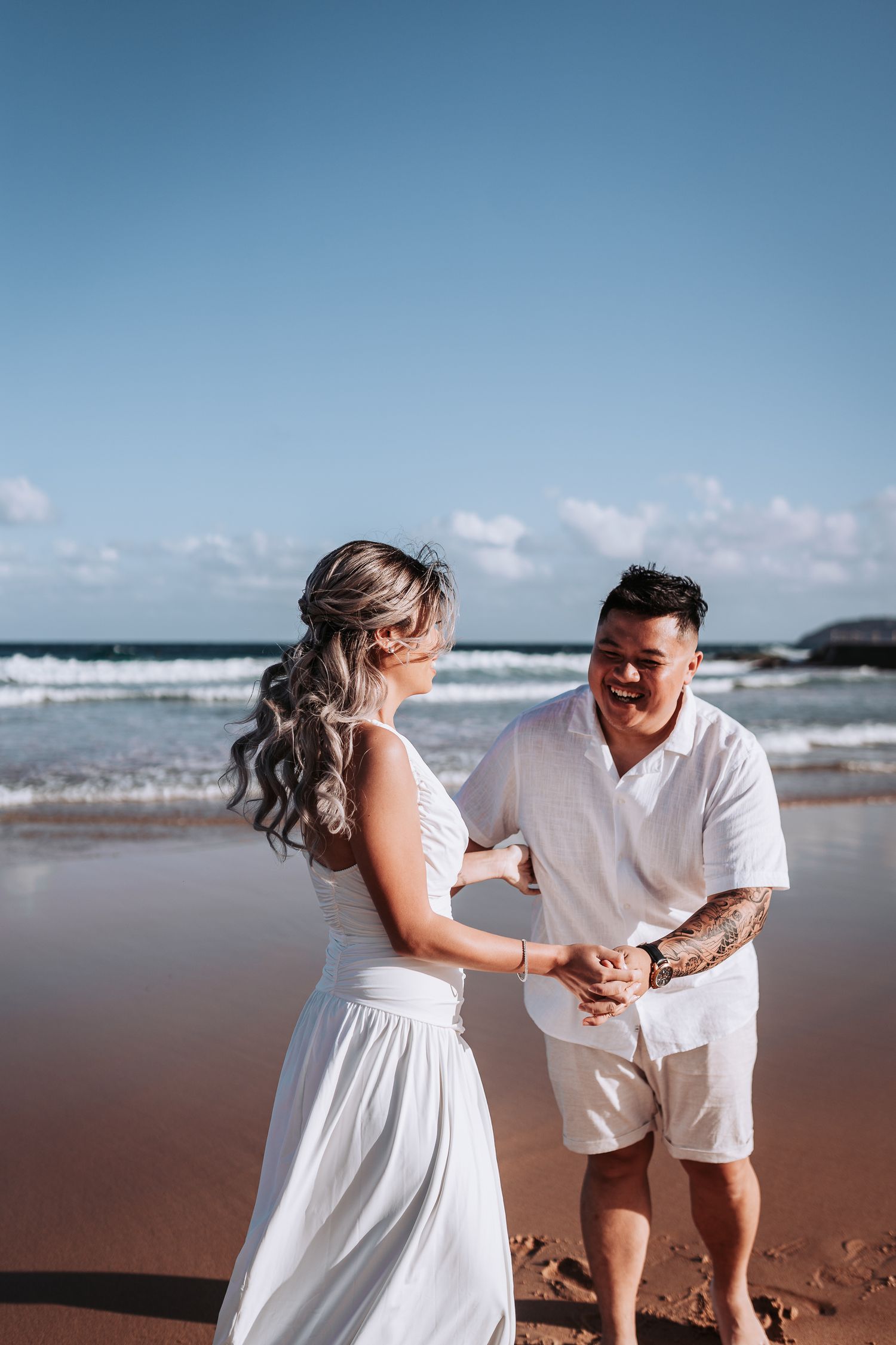 Playful beach photoshoot with couple in white outfits against ocean backdrop.