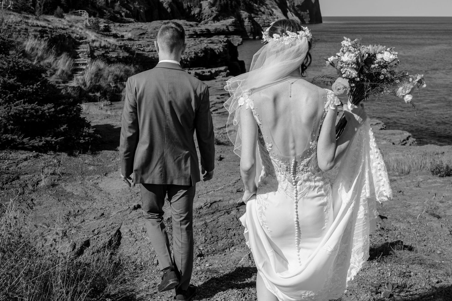 A couple on their wedding day walks along a coastal cliff path in black and white photography.