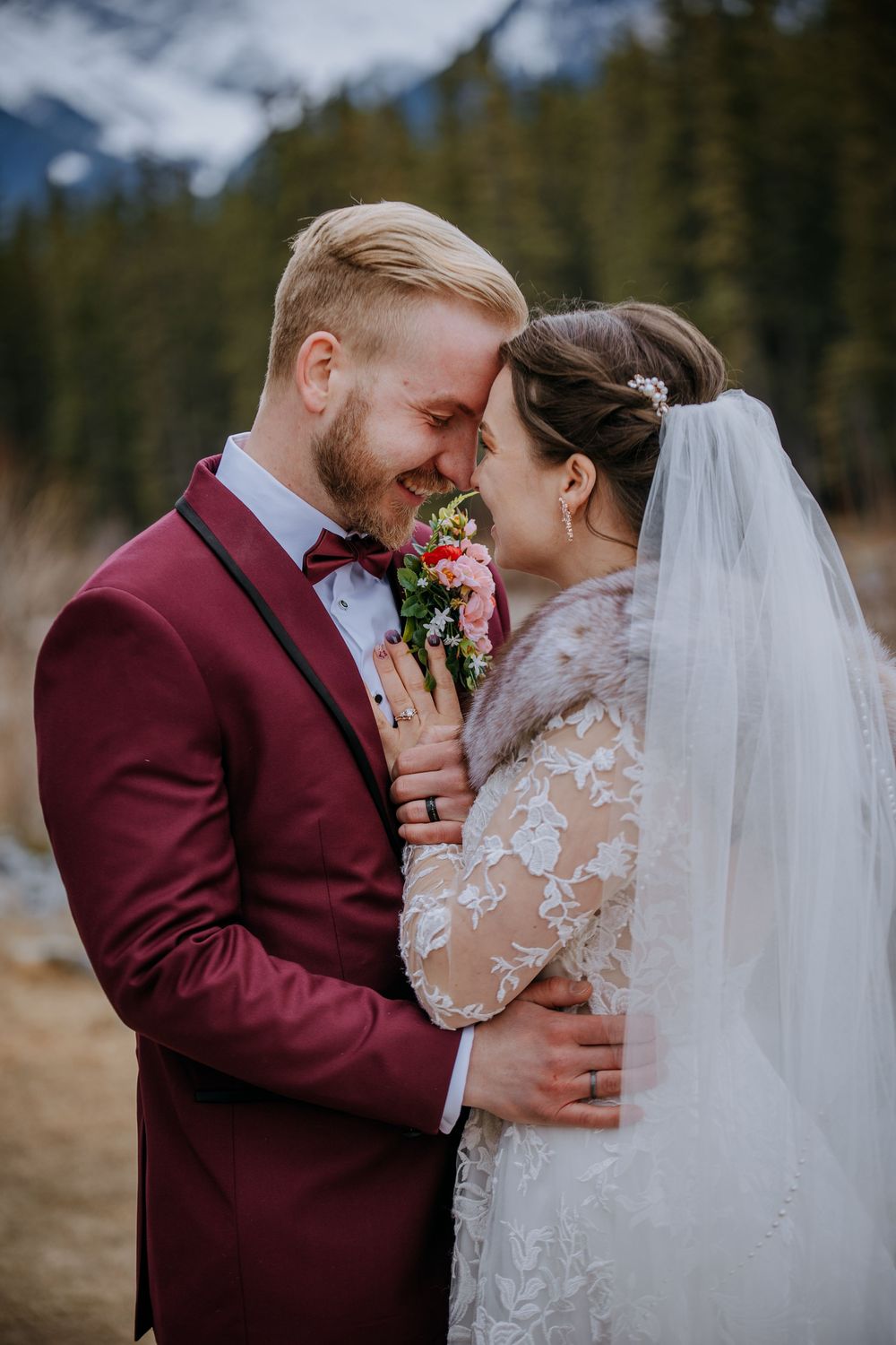A smiling groom in a burgundy suit and bride in lace dress with veil share an intimate moment outdoors with mountain backdrop.
