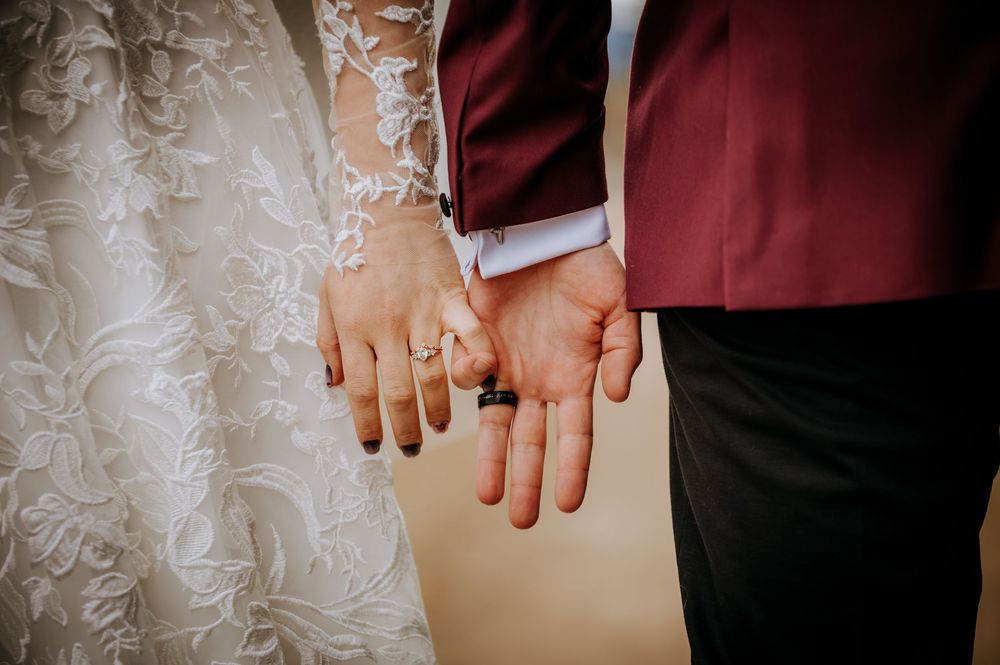 Close-up of newlyweds holding hands showing wedding rings, bride in lace gown and groom in burgundy suit.